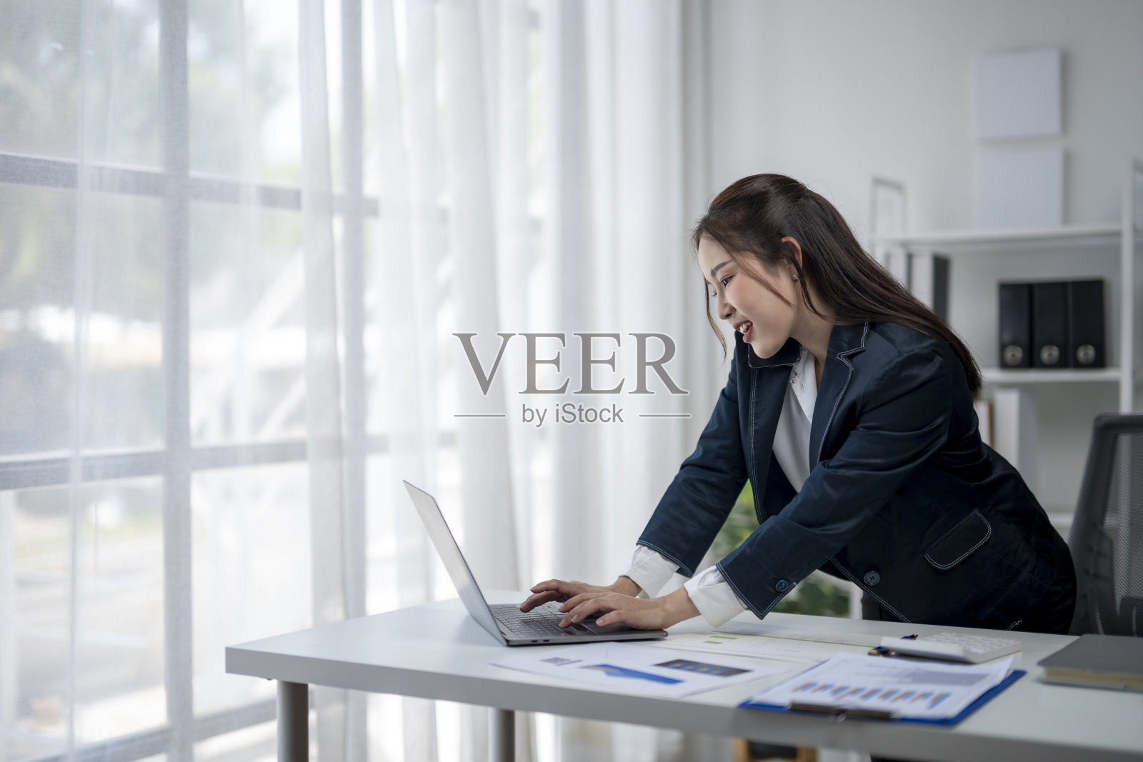 一位女士坐在桌子前，使用笔记本电脑并在打电话。她穿着西装，面带微笑。照片摄影图片