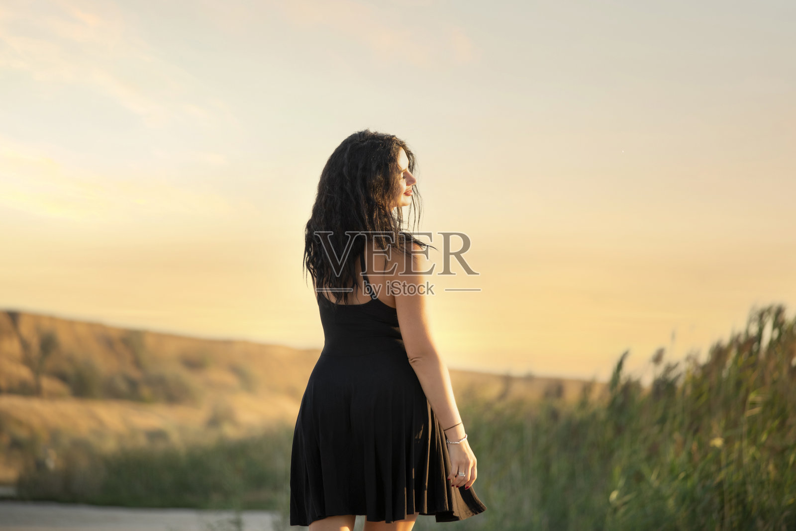 穿着短黑裙的女孩，黑色长卷发，站在野生岸边的水库岸边。照片摄影图片