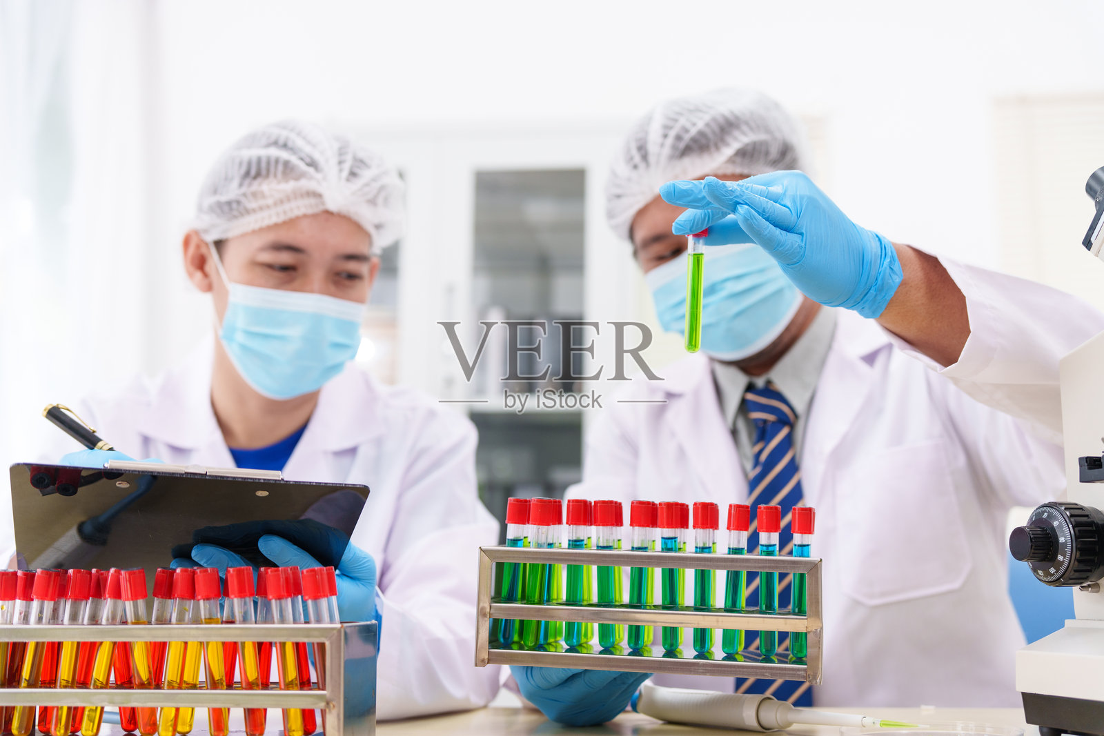 两位穿着实验室白大褂的亚洲科学家坐在实验室的桌子旁，通过显微镜检查样本。房间里配备了科学工具、液体和化学材料。照片摄影图片