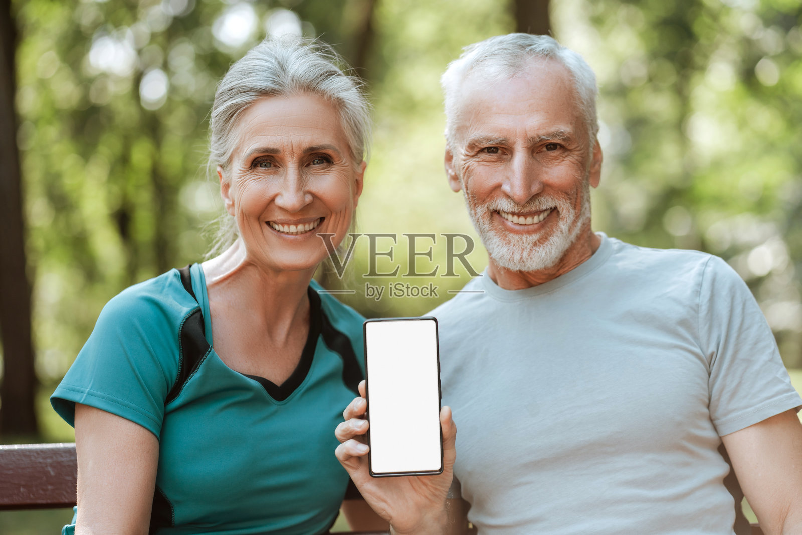 微笑的老年夫妇在公园里展示一部空白屏幕的智能手机，面向镜头。照片摄影图片