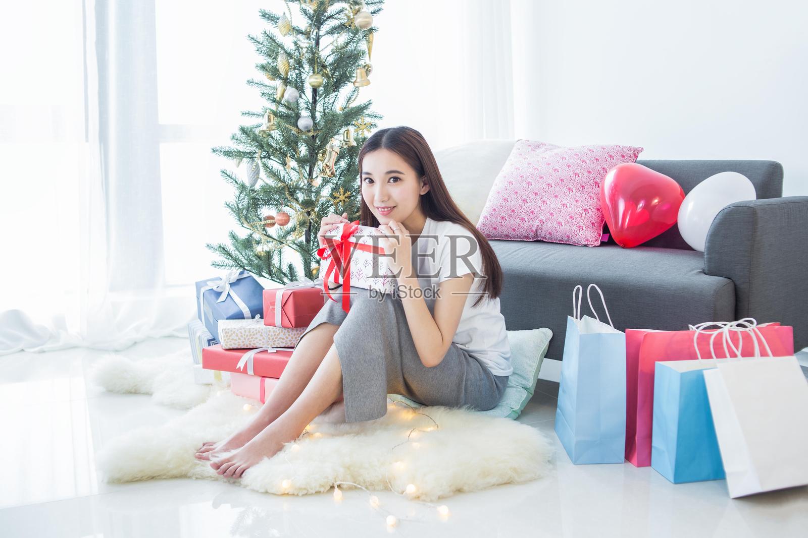 一位女性坐在圣诞树前的地板上，手里拿着一个礼物，面带微笑。照片摄影图片