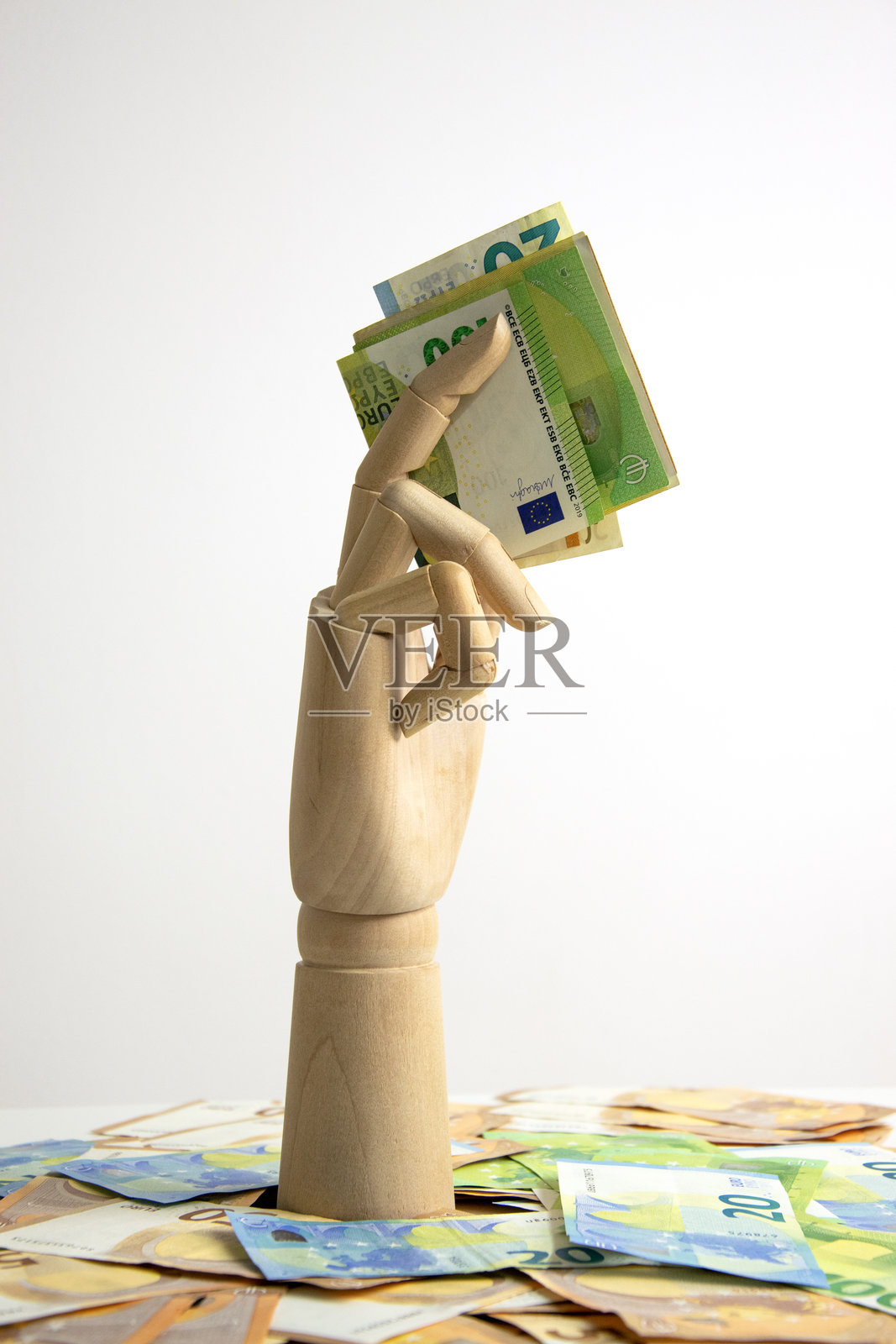 一只木制手从一堆欧元钞票中伸出，手中握着一叠欧元钞票，背景为白色。照片摄影图片