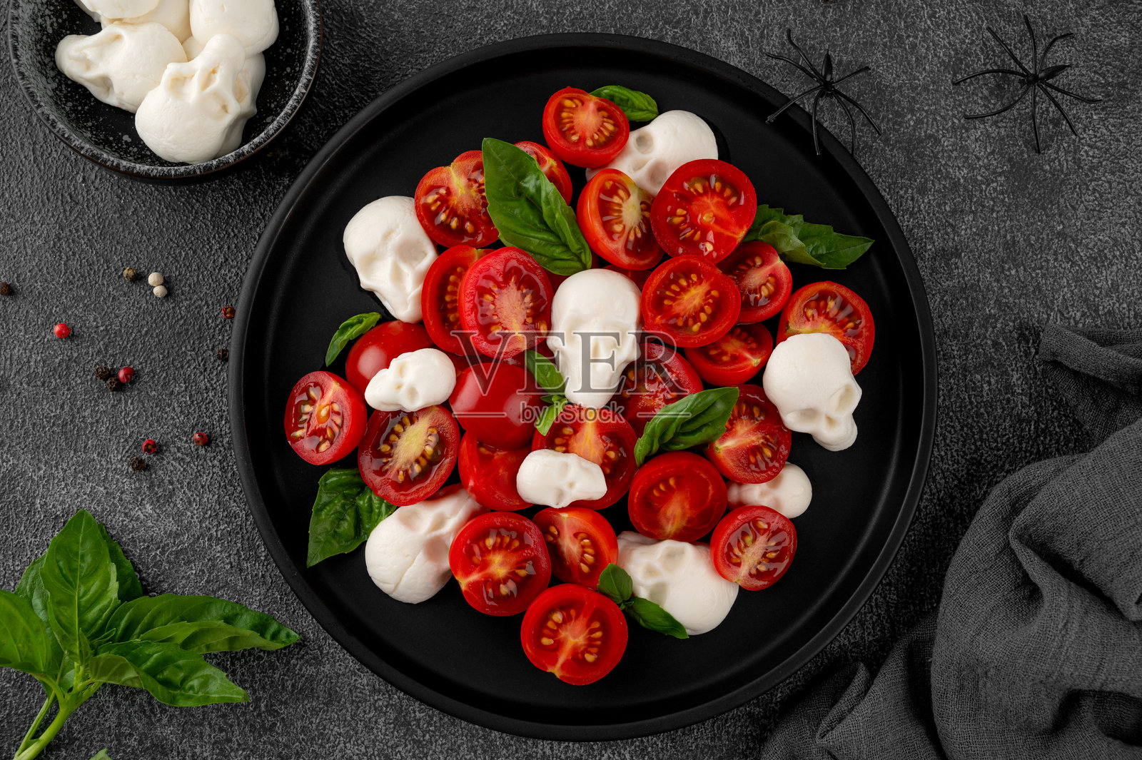 万圣节食品。黑色盘子上摆放着马苏里拉奶酪骷髅、罗勒和香醋酱的卡普雷塞沙拉，背景昏暗。搞笑的食品创意。照片摄影图片