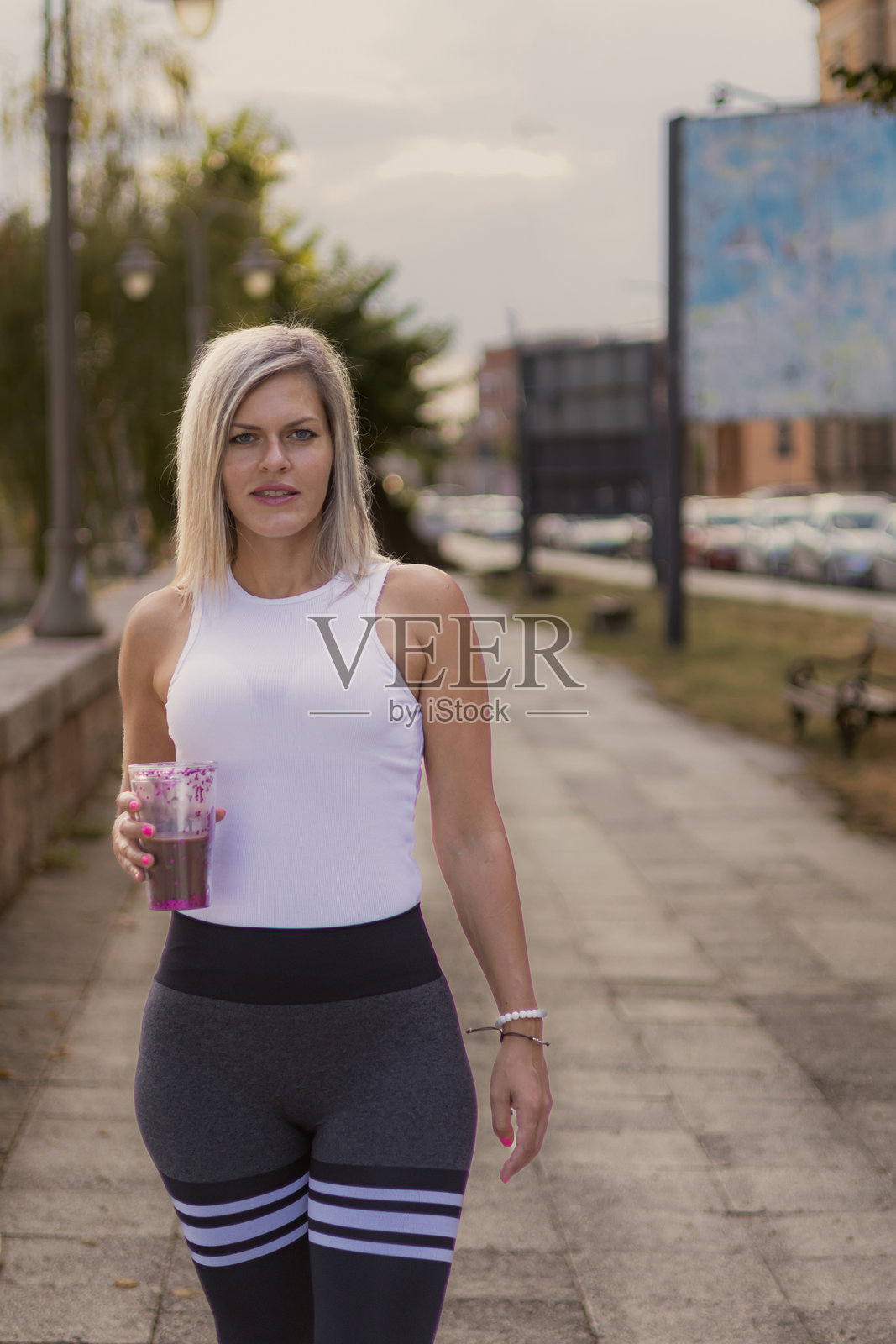 美丽的金发健身女性在公共公园户外训练后喝蛋白质奶昔。照片摄影图片