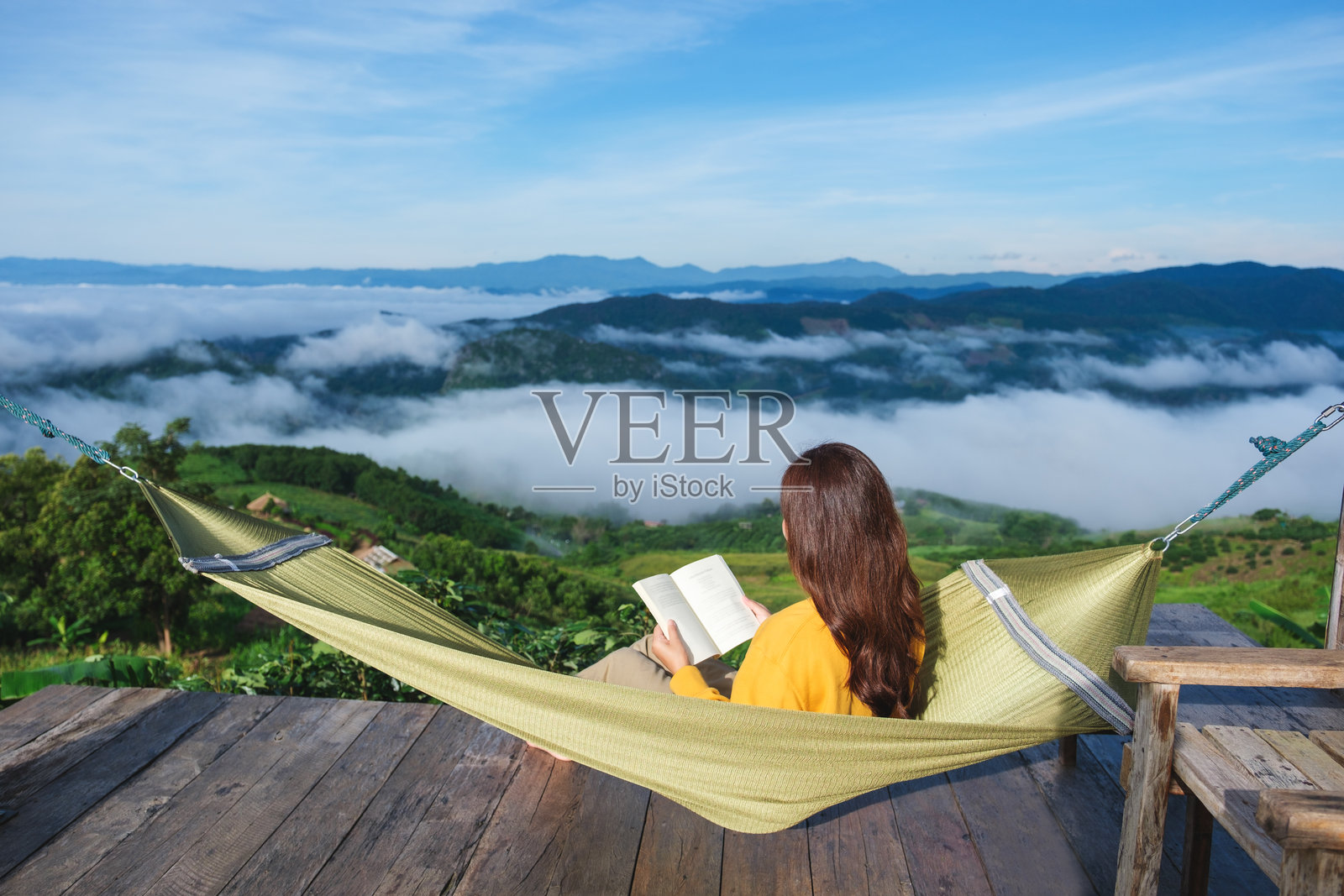 背影的女人坐在吊床上阅读书籍，背景是山景和雾海。照片摄影图片