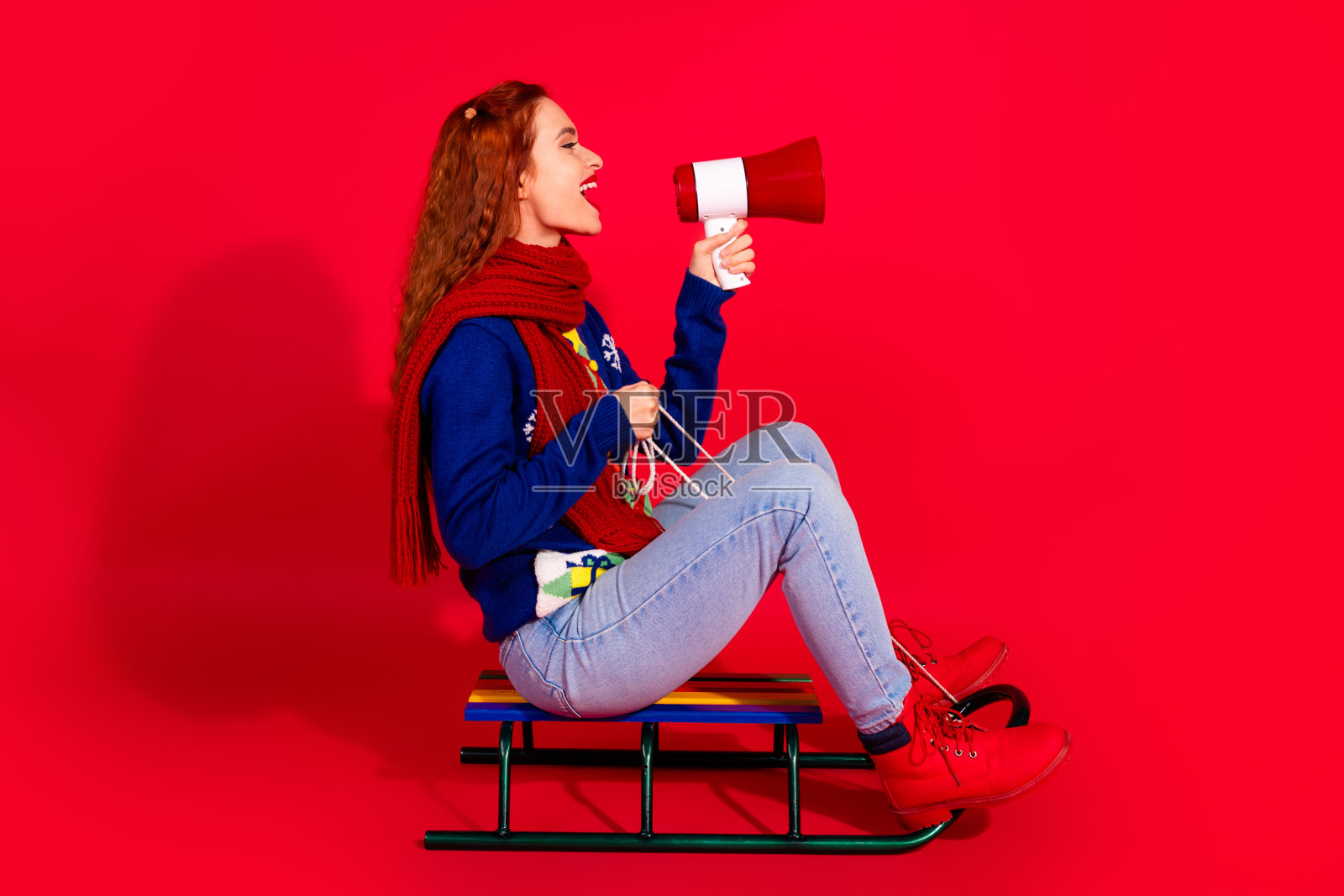 全身照的可爱年轻女士穿着蓝色圣诞服装，骑着雪橇，手持扩音器，在红色背景下庆祝冬季假期。照片摄影图片