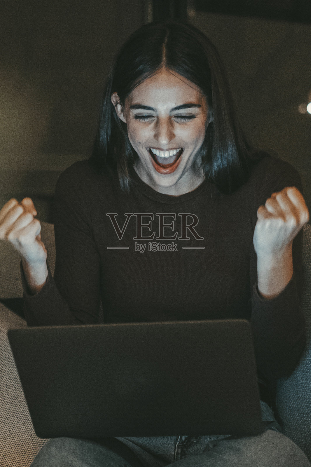 年轻女性在家中欢庆，晚上坐在沙发上举起拳头庆祝胜利，同时使用笔记本电脑。她兴奋的表情反映出成功和满足，无论是找到新工作还是伴侣。照片摄影图片