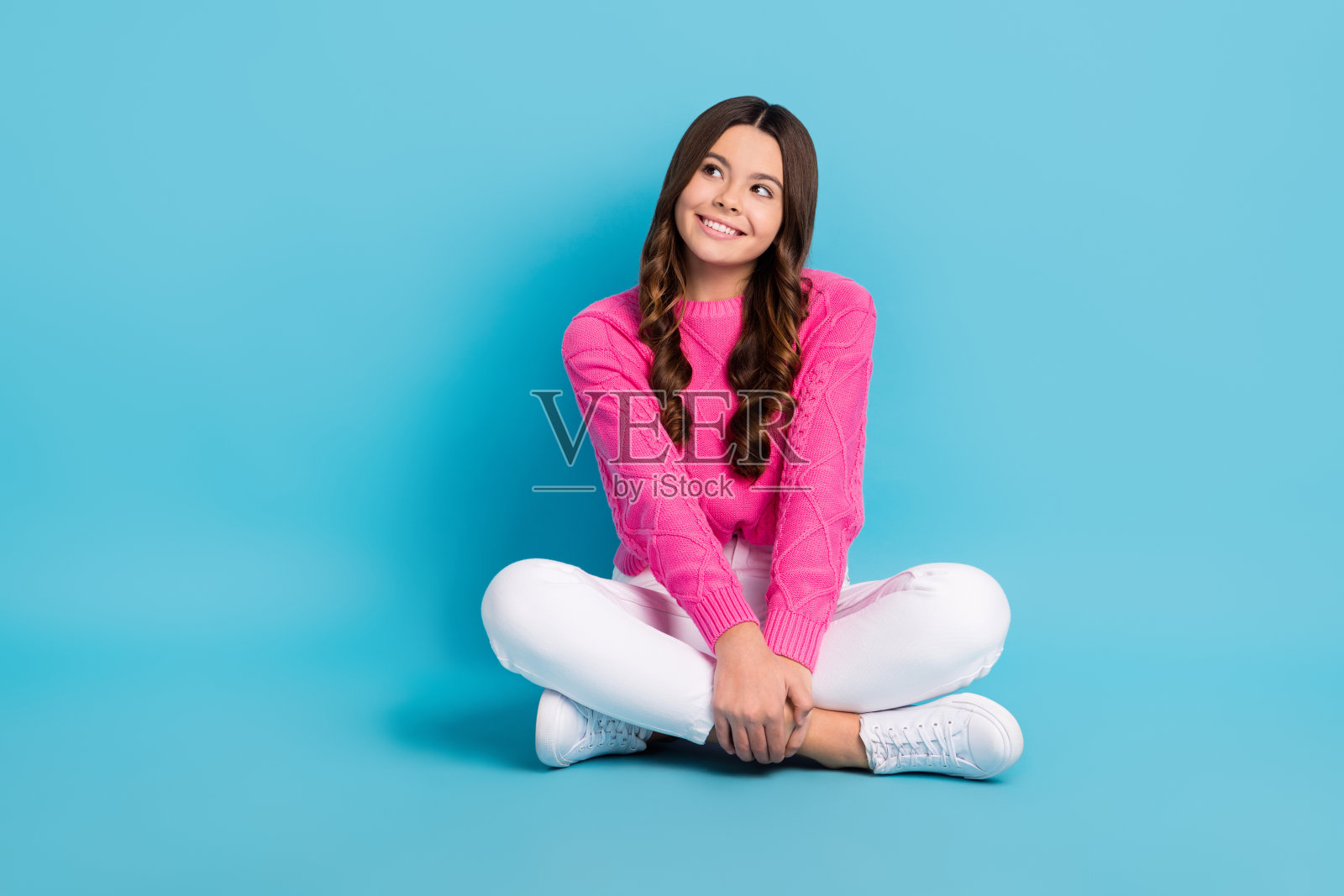 穿着粉色毛衣的梦幻甜美女子，双腿交叉坐着，凝视空旷的空间，背景为蓝色。照片摄影图片