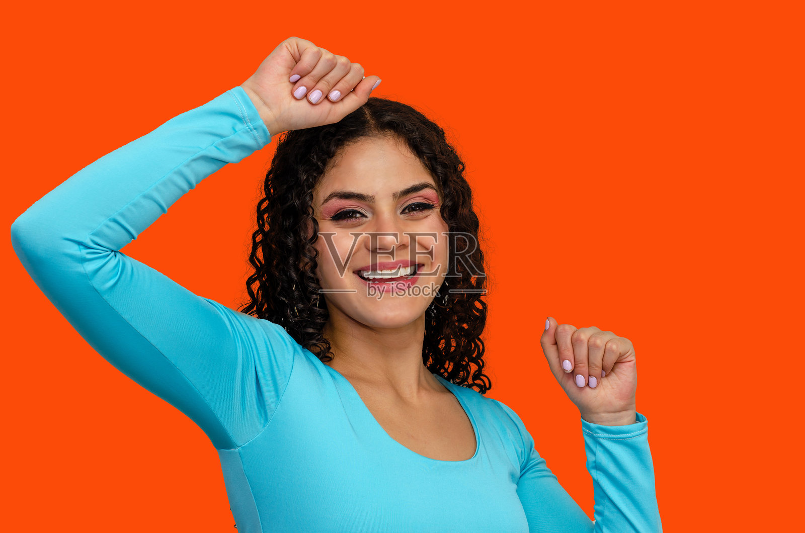 快乐美丽的年轻女性特写肖像，卷发，穿着色彩斑斓的休闲服，双臂高举，背景为橙色。照片摄影图片