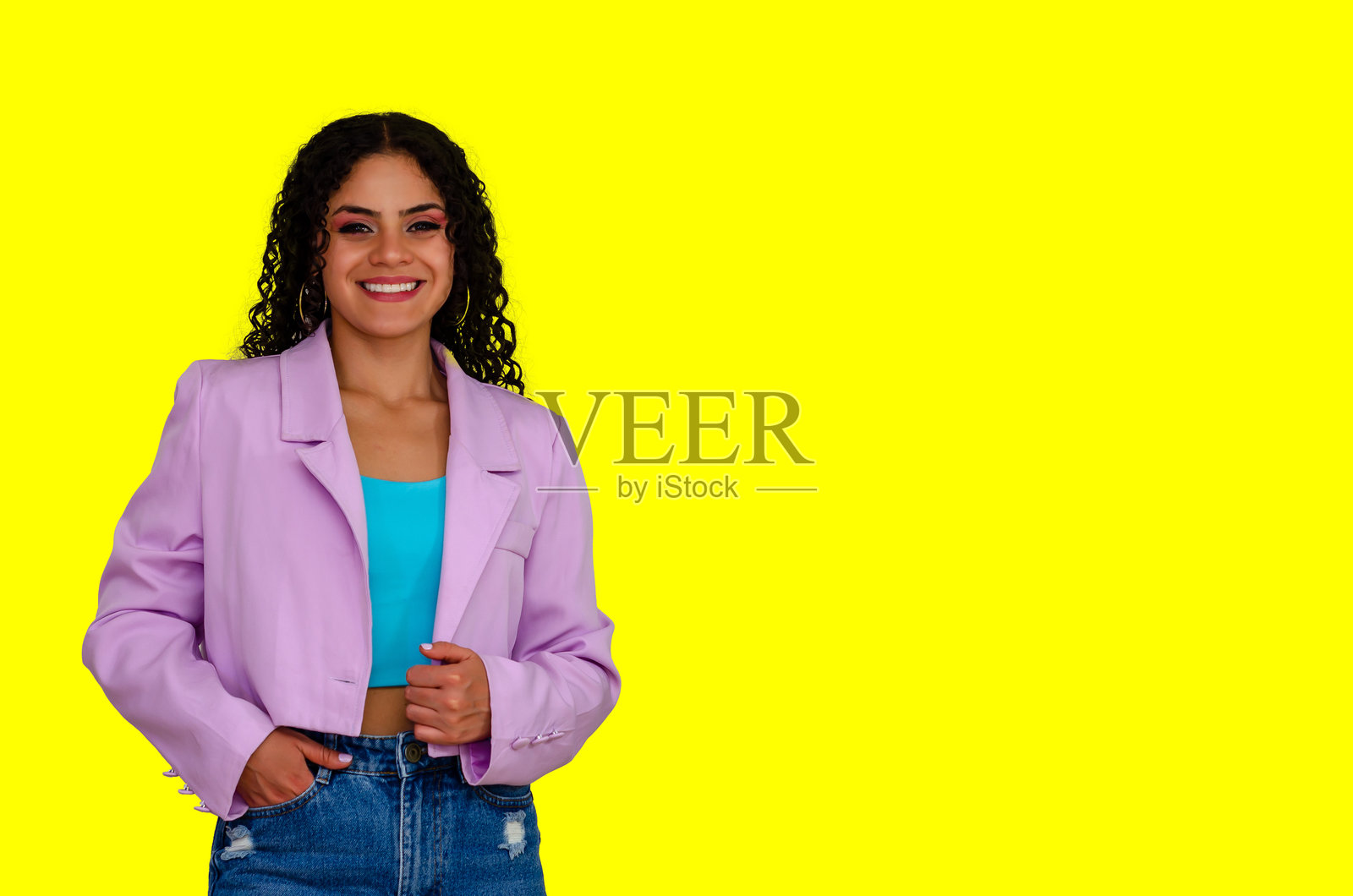 一位快乐美丽的年轻女性，卷发，穿着色彩斑斓的休闲服，双手插在口袋里，背景为黄色。照片摄影图片