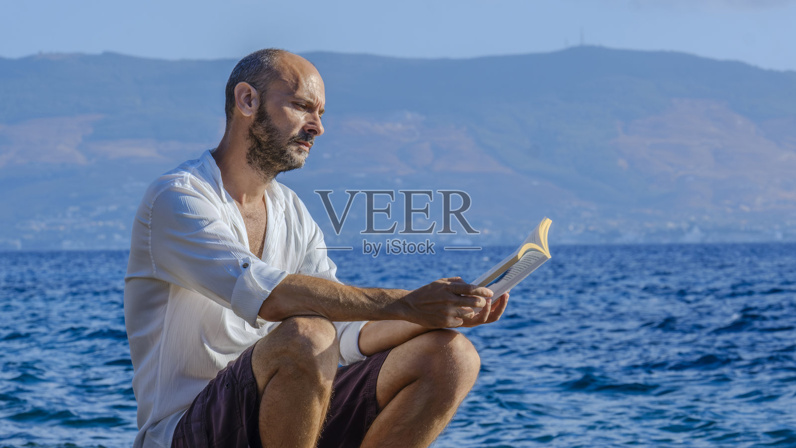 一位中年魅力男士穿着白衬衫，坐在海边的岩石上，在阳光明媚的夏日里阅读一本书，享受阳光，度假放松。照片摄影图片