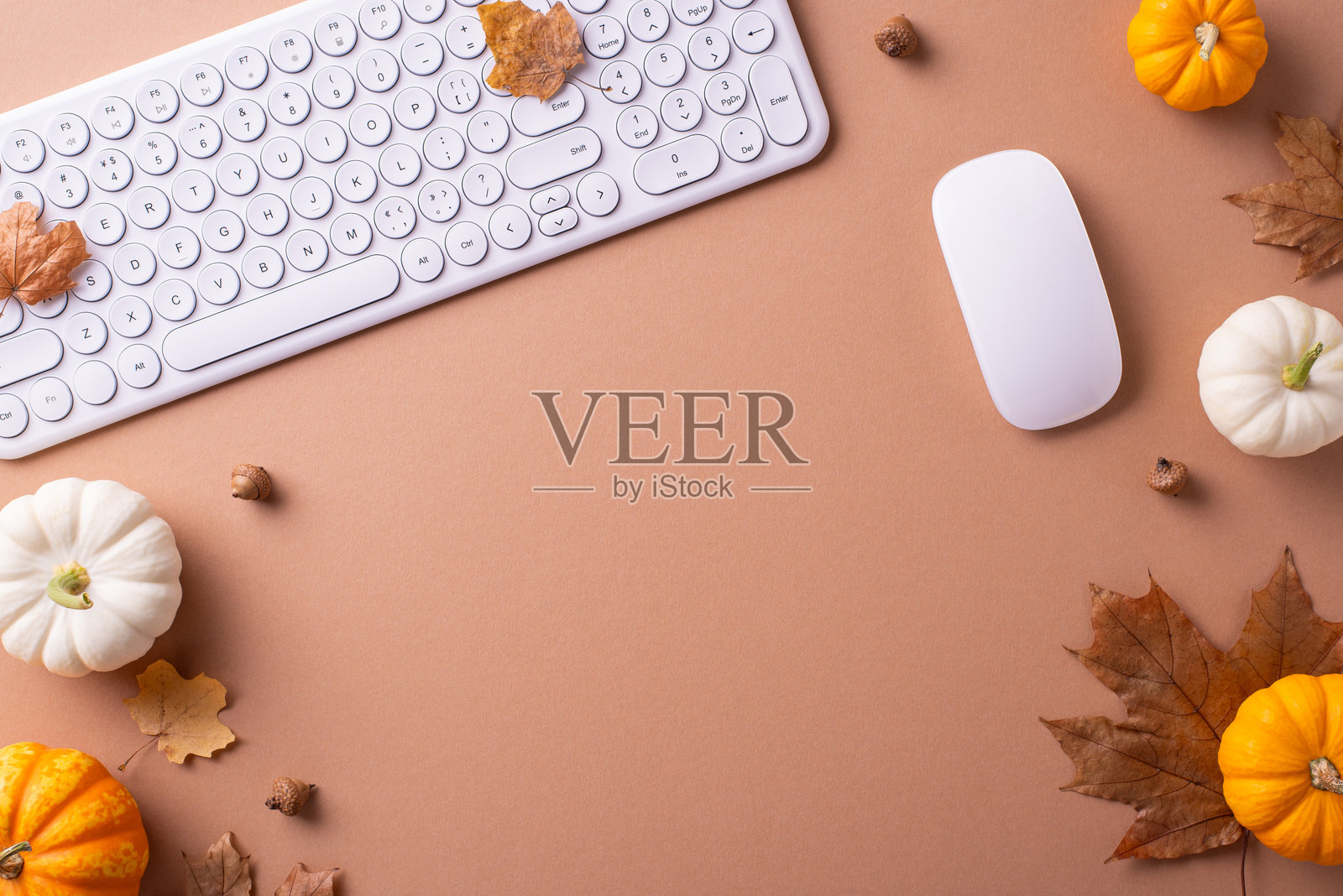 米色工作空间的平铺图，白色键盘和鼠标被南瓜和秋叶环绕，营造出温馨的秋季氛围照片摄影图片