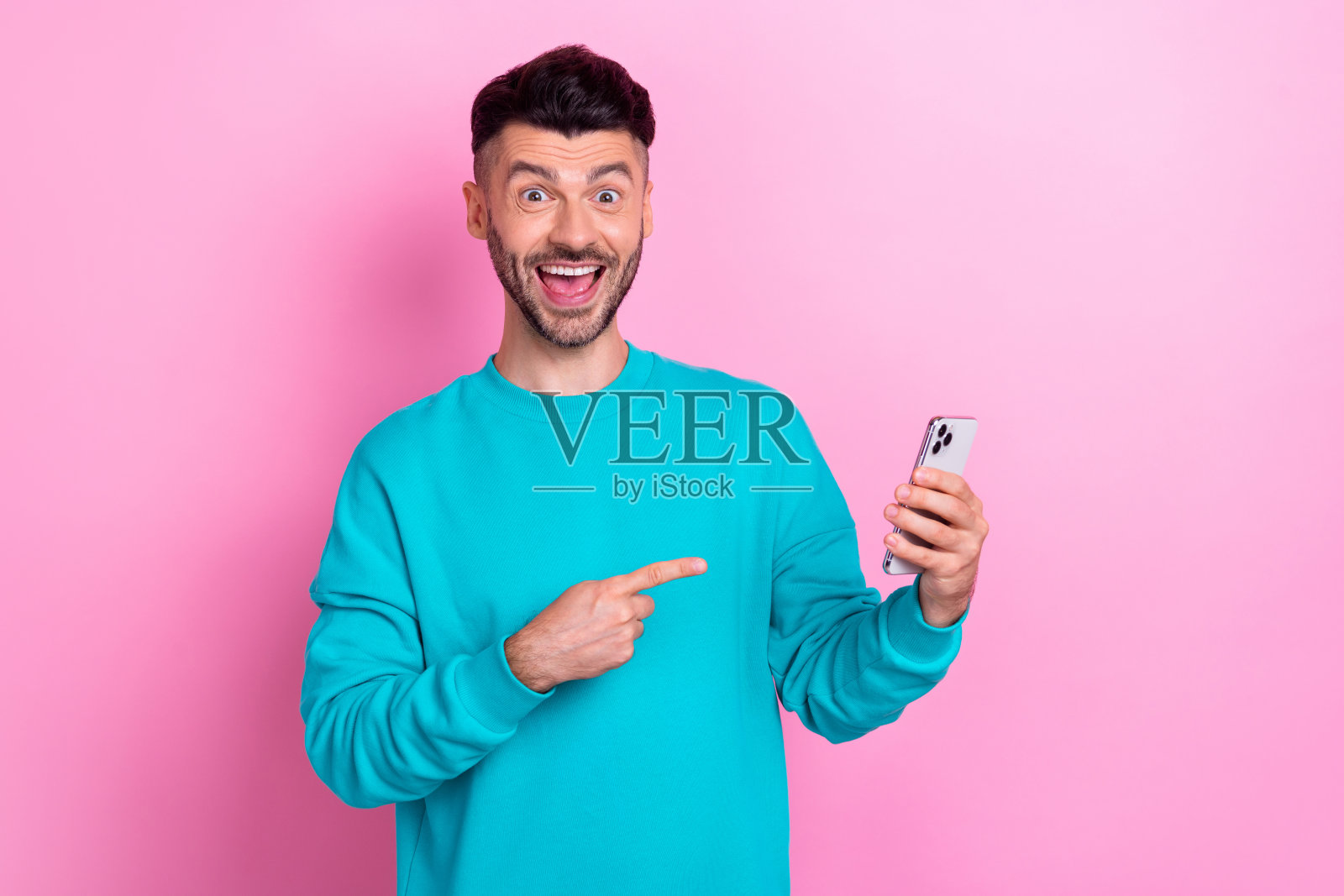 惊讶而快乐的男士肖像，手指直接指向手持电话，背景为粉色。照片摄影图片