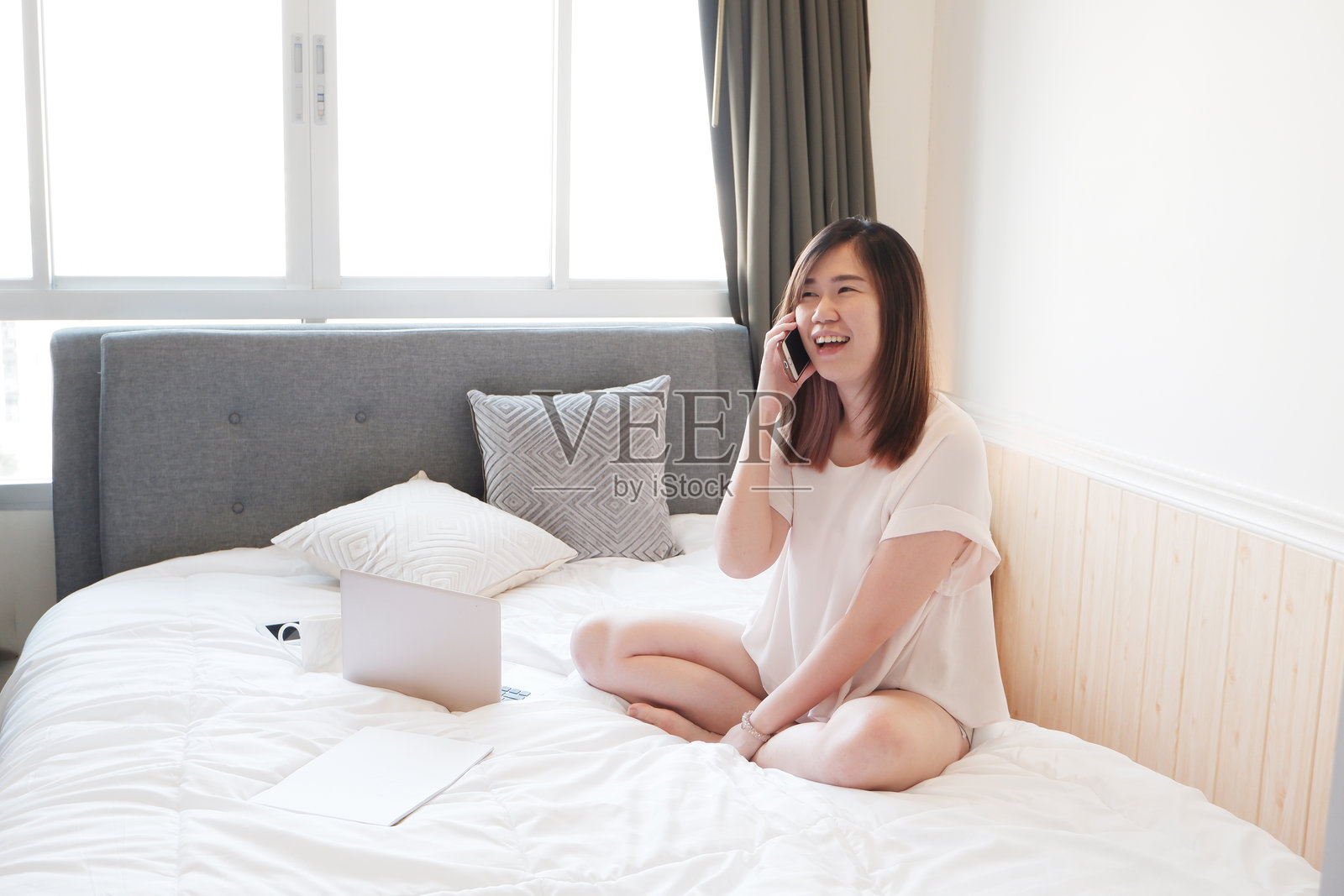 微笑的亚洲年轻女性在卧室的床上放松，使用智能手机交谈。照片摄影图片