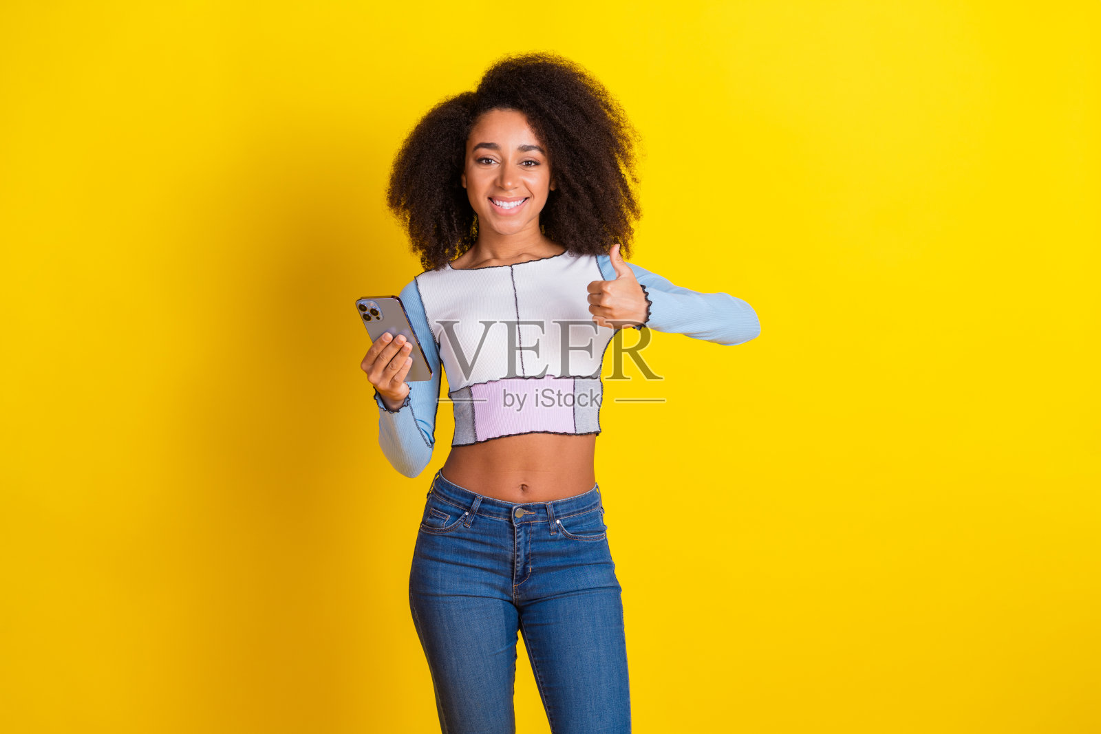 可爱的年轻女士在黄色背景下穿着时尚的蓝色服装，手持设备竖起大拇指的照片肖像照片摄影图片