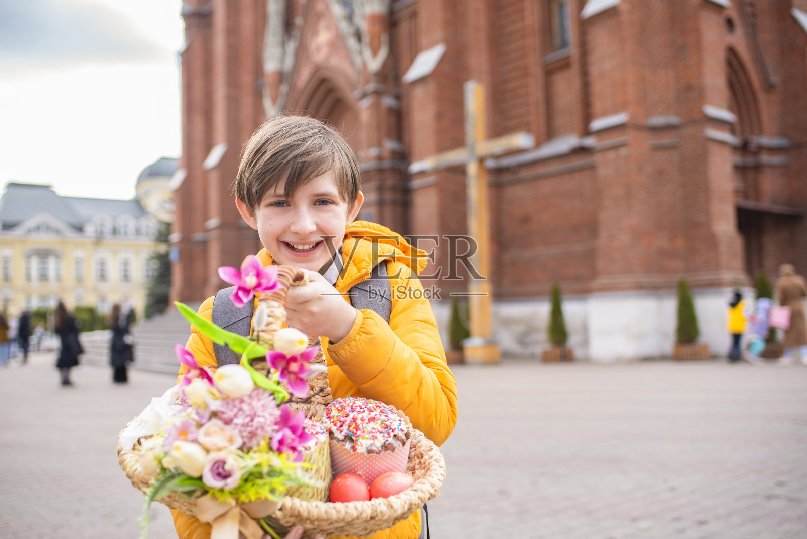 在一座砖砌教堂附近，一个面带微笑的11岁男孩拿着一篮子传统的复活节蛋糕、巧克力和彩蛋。照片摄影图片