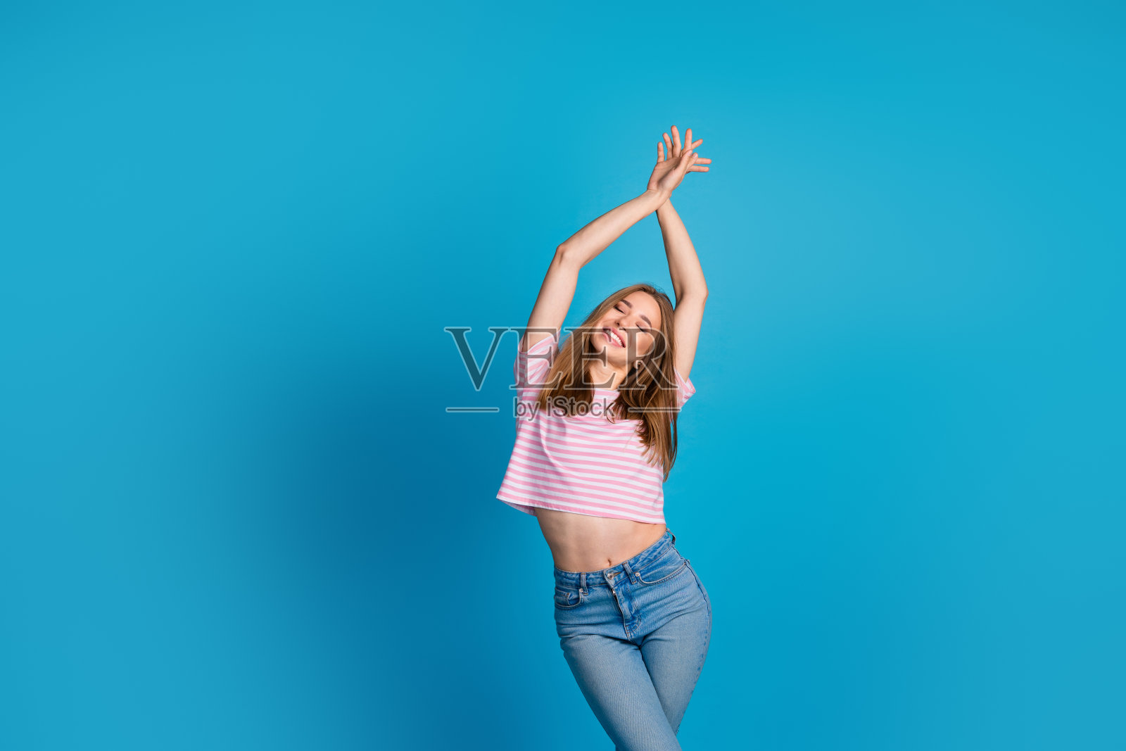 可爱年轻女士在蓝色背景下优雅地舞动，双手举起，穿着时尚的粉色条纹服装。照片摄影图片
