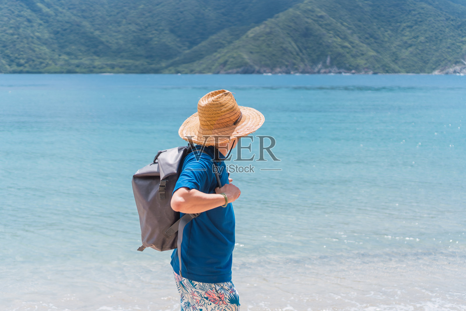 戴着草帽和背包的男人在海滩边行走，远处是山照片摄影图片