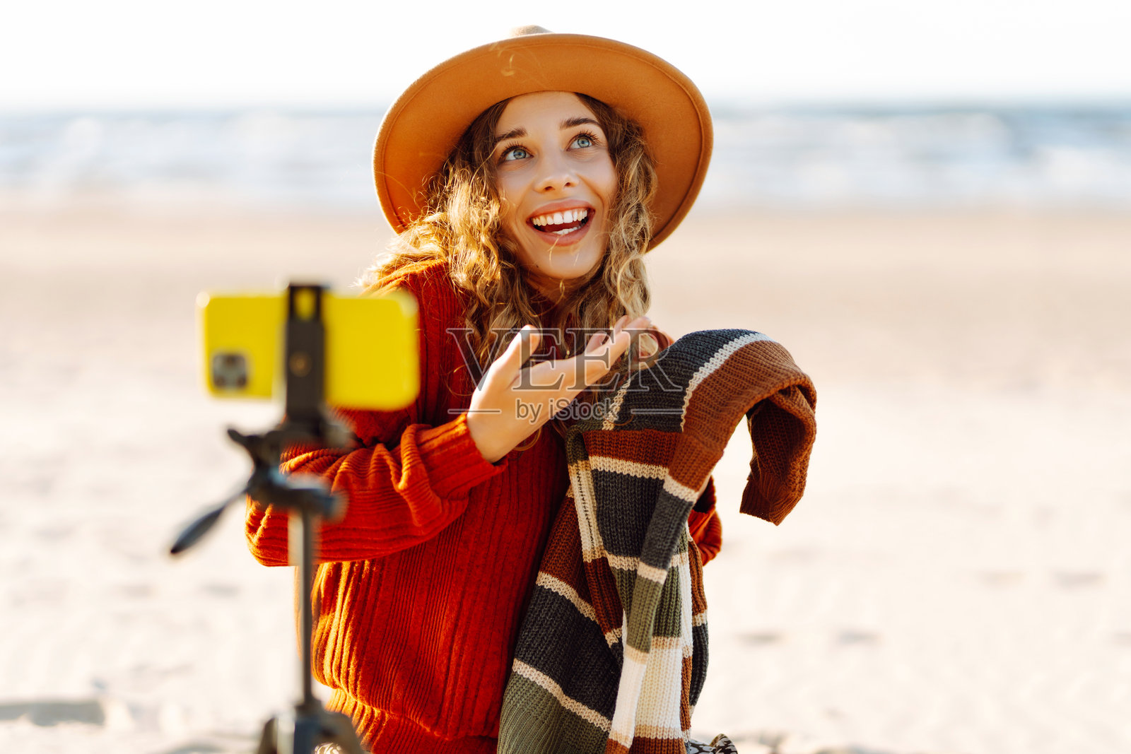 一位快乐的女性博主，卷发微波，面带灿烂笑容，在沙滩上录制视频。照片摄影图片