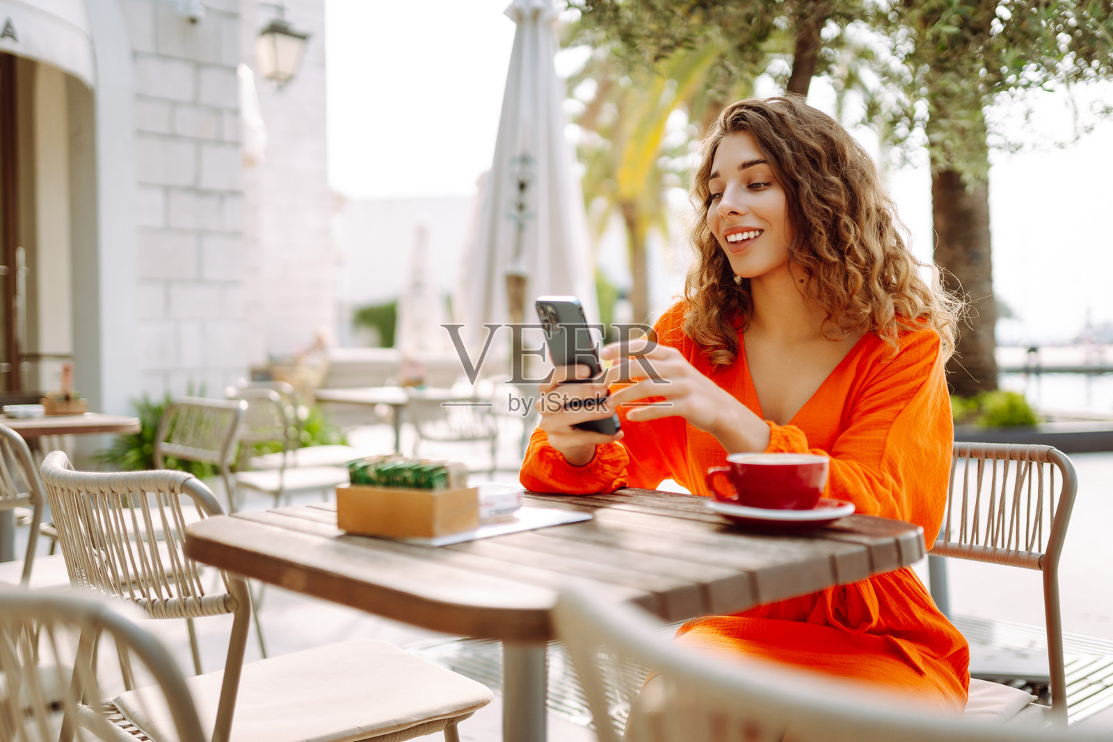 一位戴着时尚太阳镜的女性在阳光明媚的咖啡馆中享受咖啡，同时使用她的智能手机，周围环绕着棕榈树。照片摄影图片