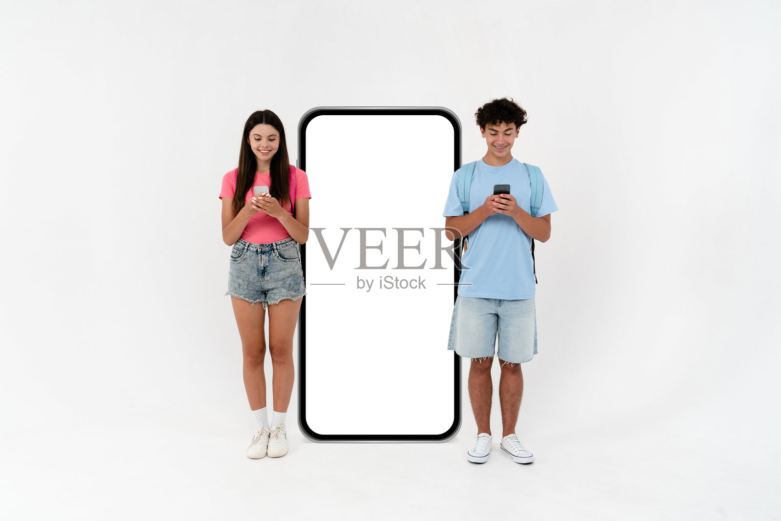 一群高中生男孩女孩穿着休闲服，背着书包，站在一个巨大的空白屏幕前，手持手机，适合在白色背景下使用的工作空间。照片摄影图片