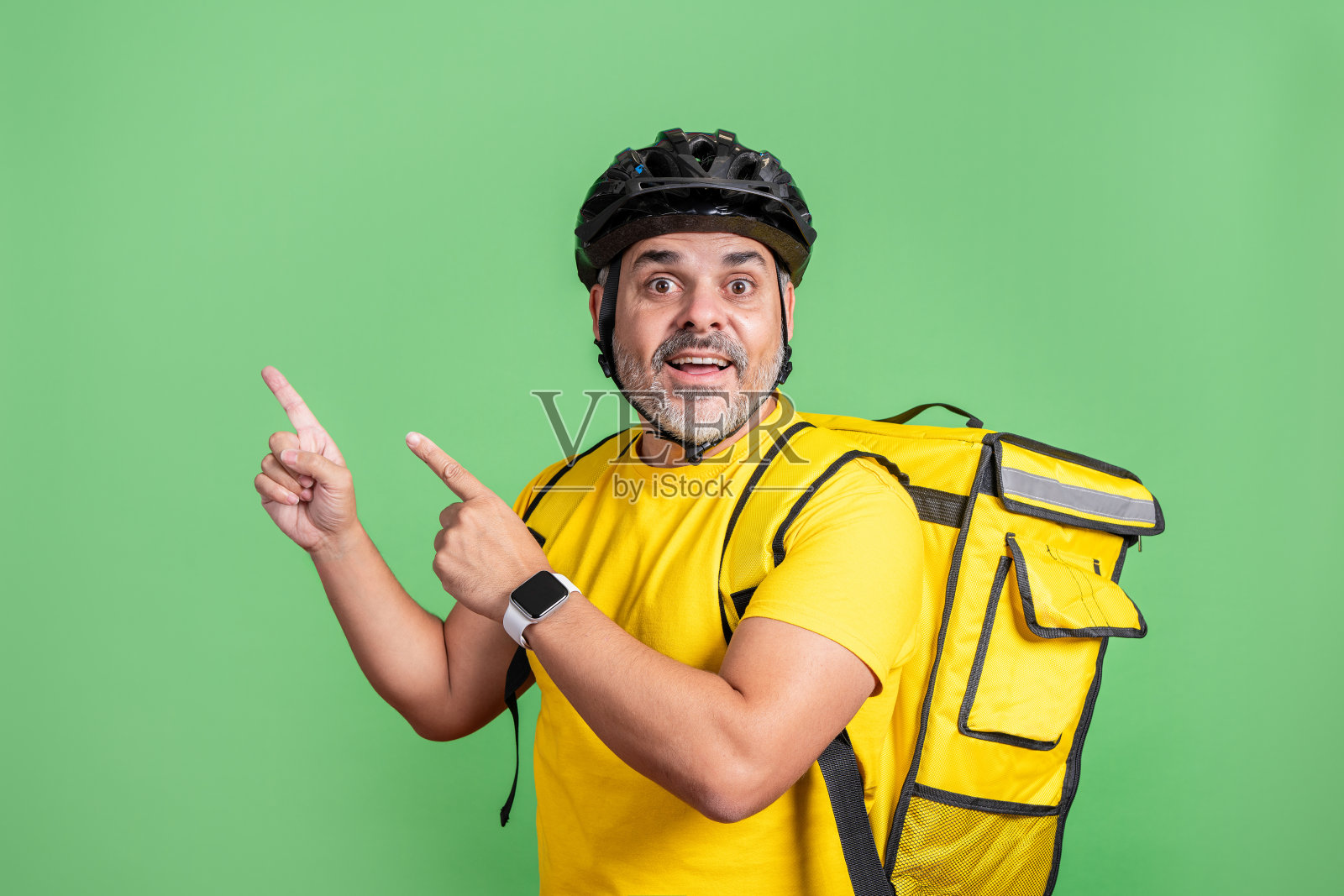 身穿黄色的快递员在绿色背景下兴奋地指着，头戴安全帽。照片摄影图片