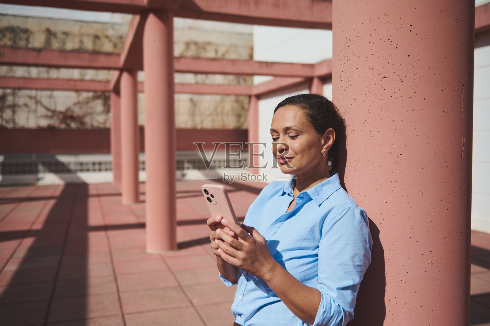 阳光明媚的日子里，女人靠在柱子上放松，使用智能手机。照片摄影图片