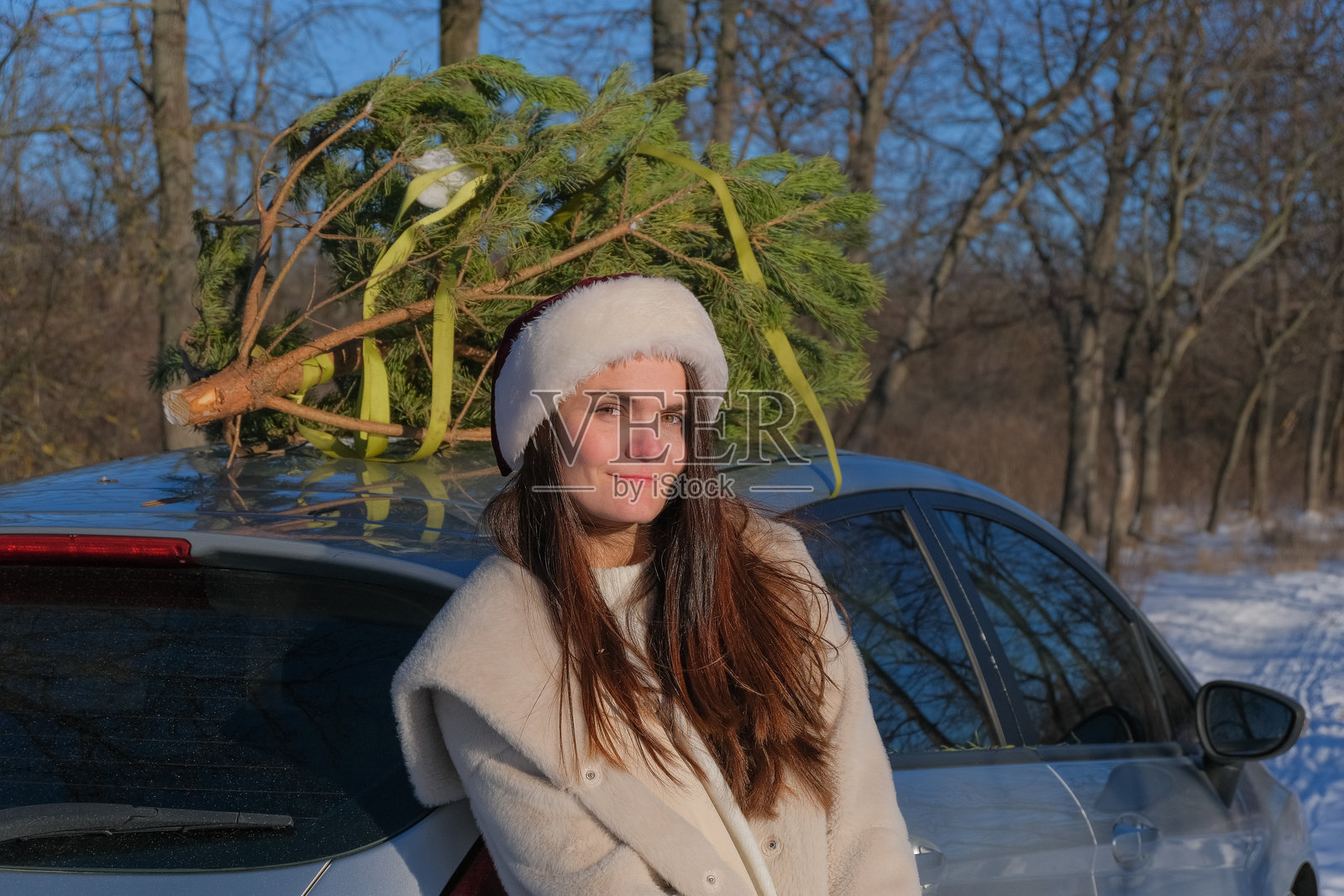 年轻女性戴着圣诞老人帽，靠在车上，车顶上放着圣诞树照片摄影图片