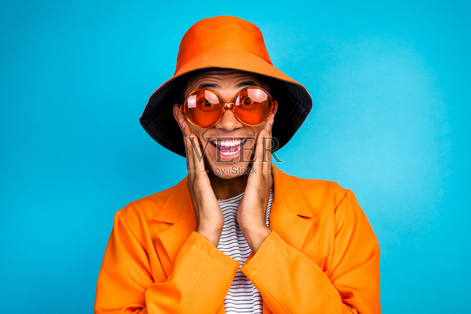 穿着时尚潮流服装的积极男士，触摸脸庞，感到印象深刻，背景为蓝色。照片摄影图片