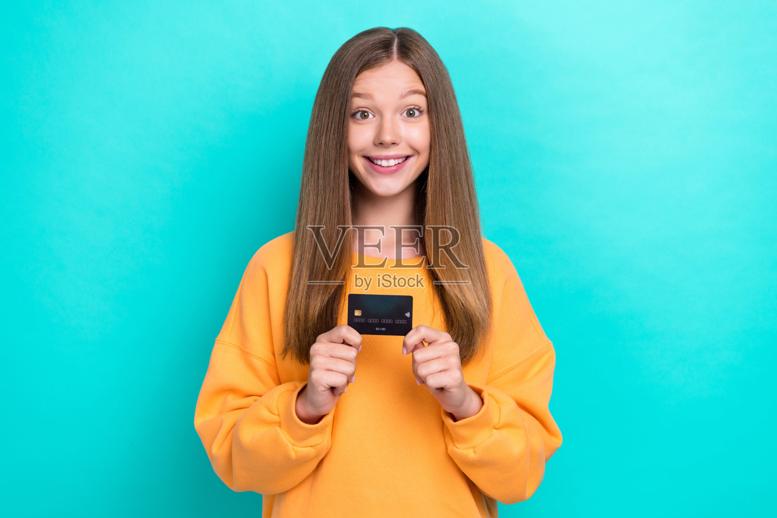 兴奋的女孩青少年穿着时尚毛衣，手持塑料信用卡，想要在周末花钱娱乐，背景为青色。照片摄影图片