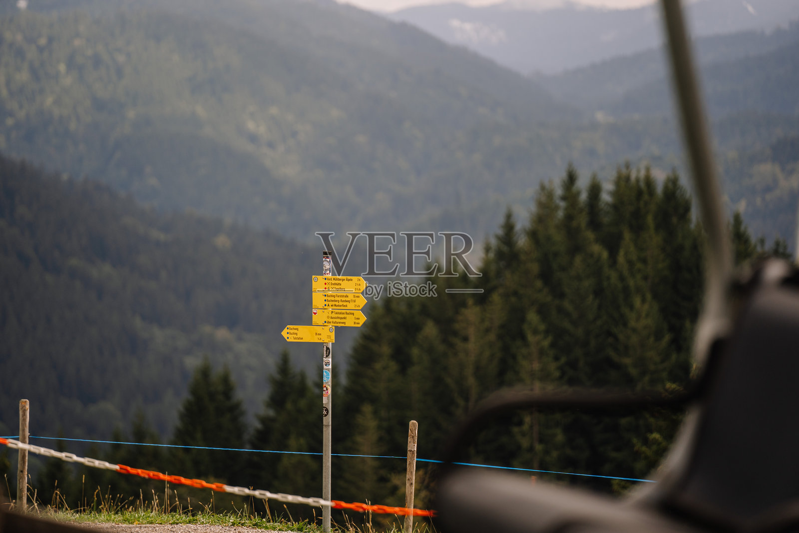 一根黄色的登山指示牌矗立在巴伐利亚的山地风景中，周围是树木和朦胧的山丘。照片摄影图片