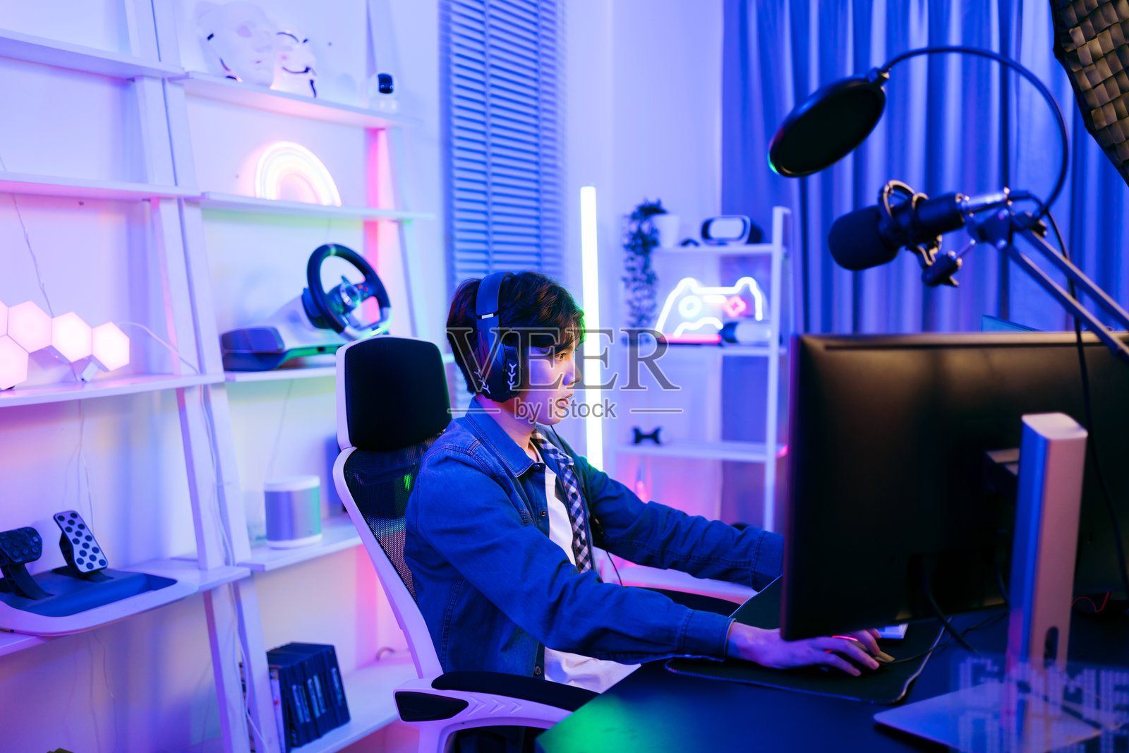 一位年轻的亚洲男性职业玩家穿着牛仔夹克，坐在椅子上，面前是一个配有电脑、键盘、显示器和麦克风的游戏桌。他正在为比赛做准备，进行游戏直播或录制播客。照片摄影图片