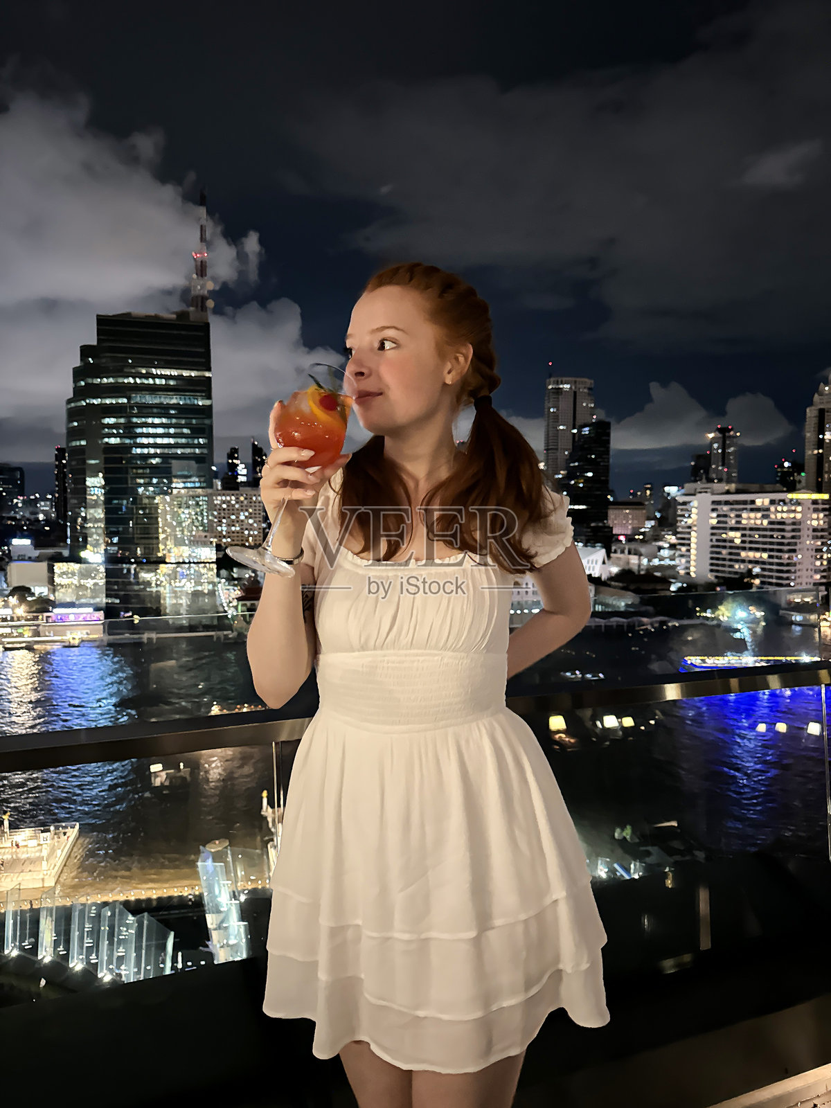 迷人的红发女子身穿挤奶女工款式的连衣裙，站在阳台玻璃安全护栏后面，手持一杯鸡尾酒，背景是曼谷湄南河上的摩天大楼天际线，以及在暮色中被照亮的建筑物照片摄影图片