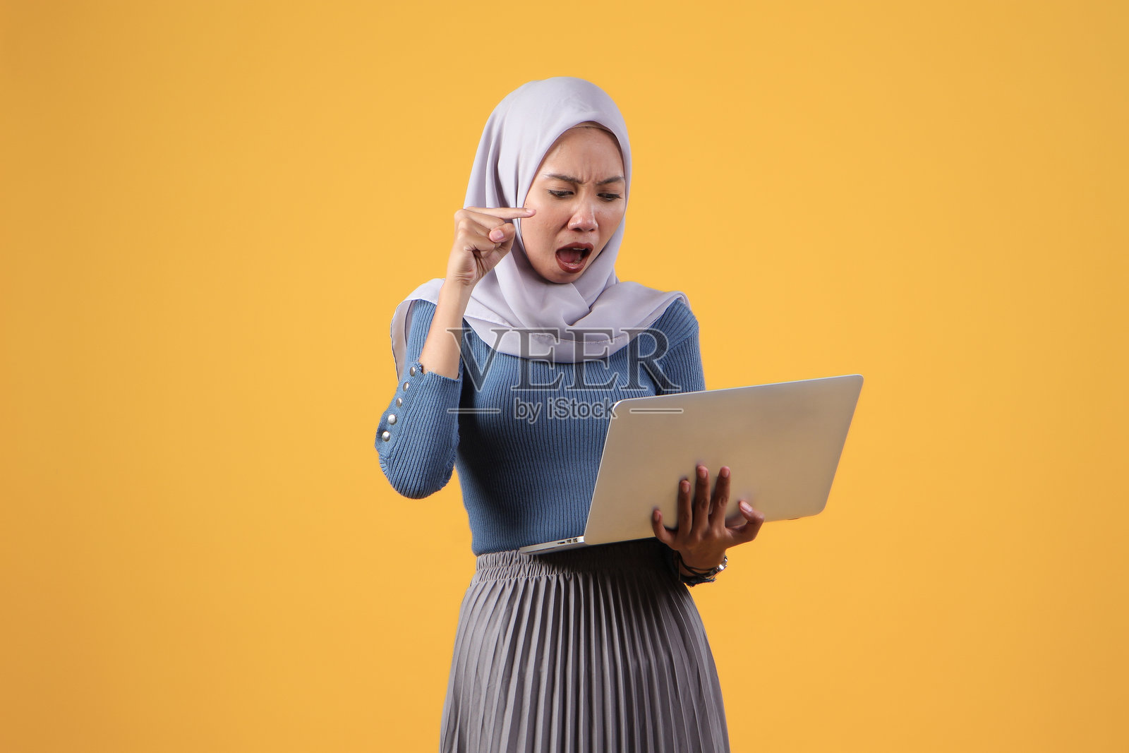 在黄色背景上拿着笔记本电脑、面带怒容的印尼穆斯林妇女照片摄影图片