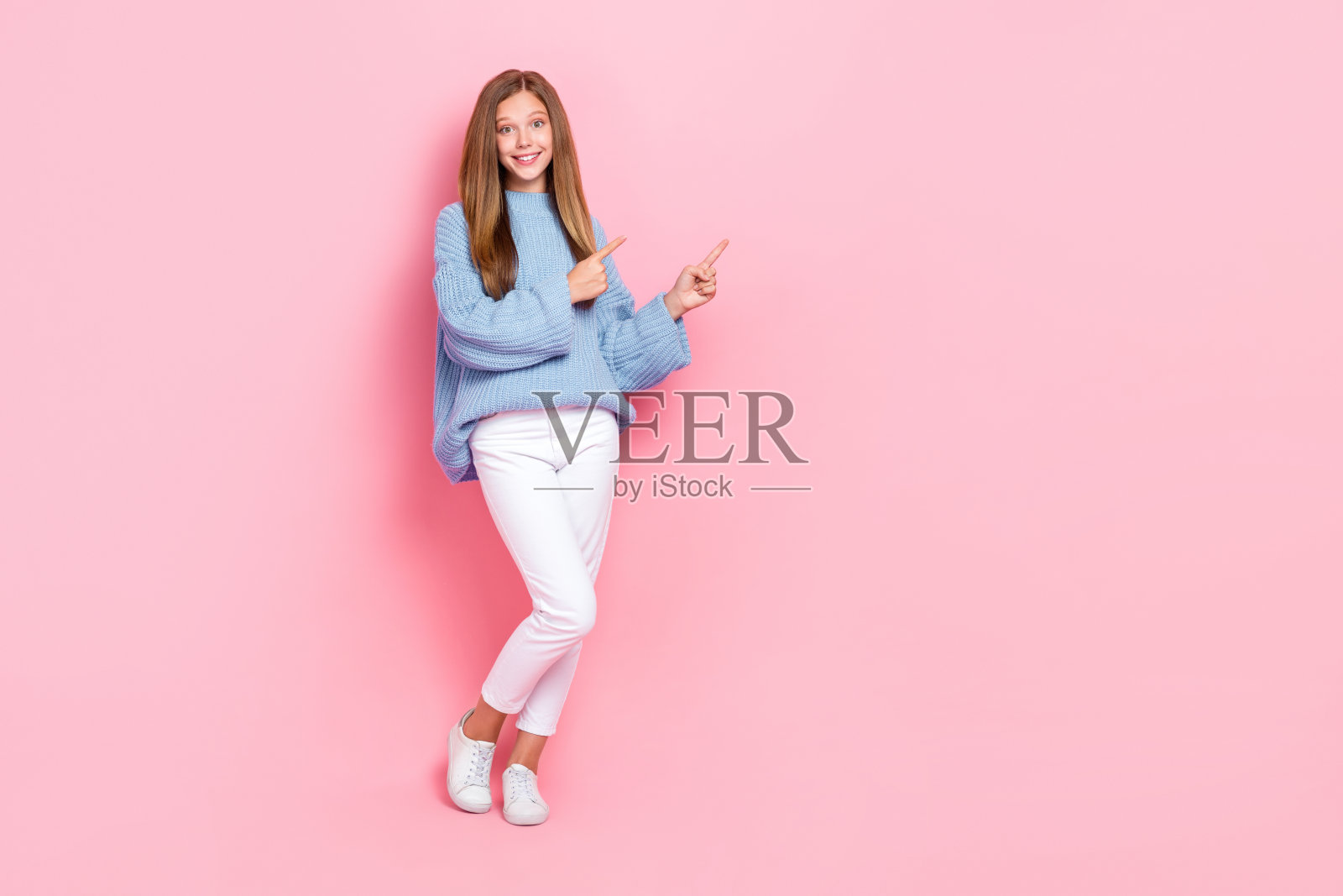 可爱的年轻女学生全身照，穿着时尚服装，手指指向空白处，展示品牌购物，背景为粉色。照片摄影图片