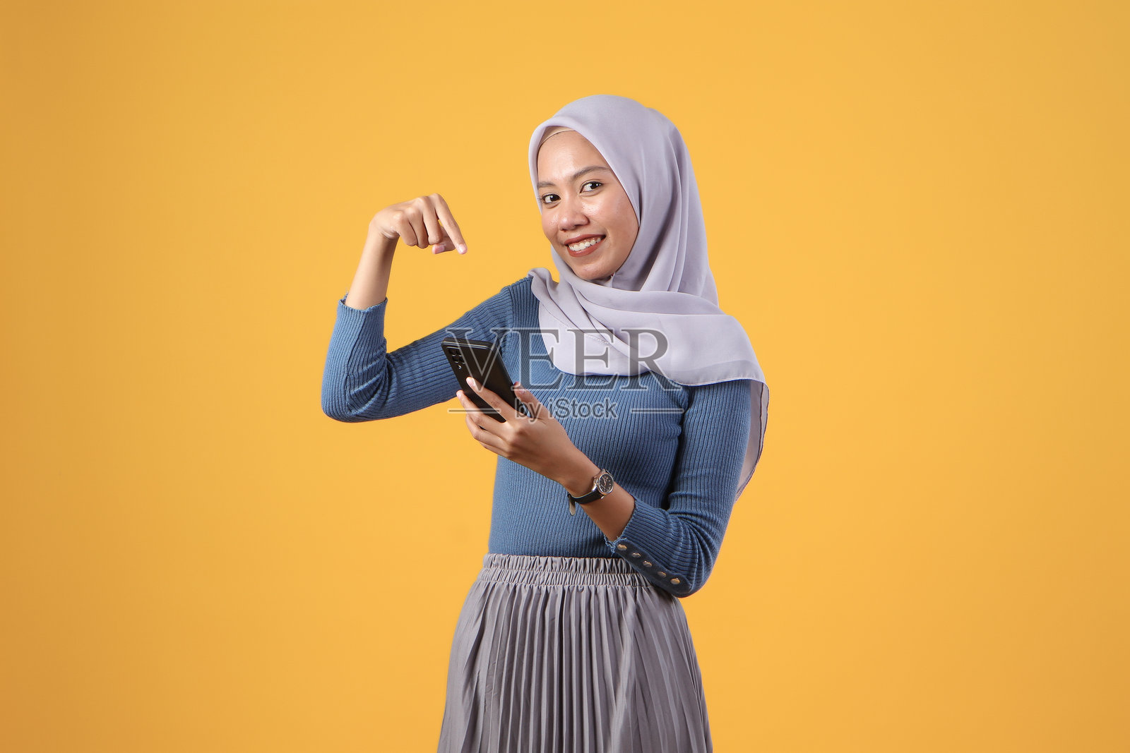 快乐的印尼穆斯林妇女在黄色背景下手持智能手机并指向前方照片摄影图片