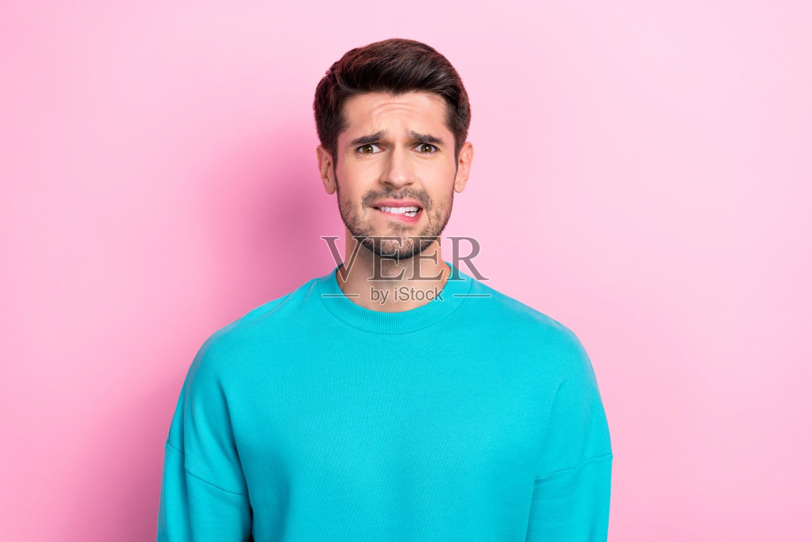 时尚蓝色着装的帅气年轻男子，咬着嘴唇，神情困惑和担忧，背景为粉色。照片摄影图片