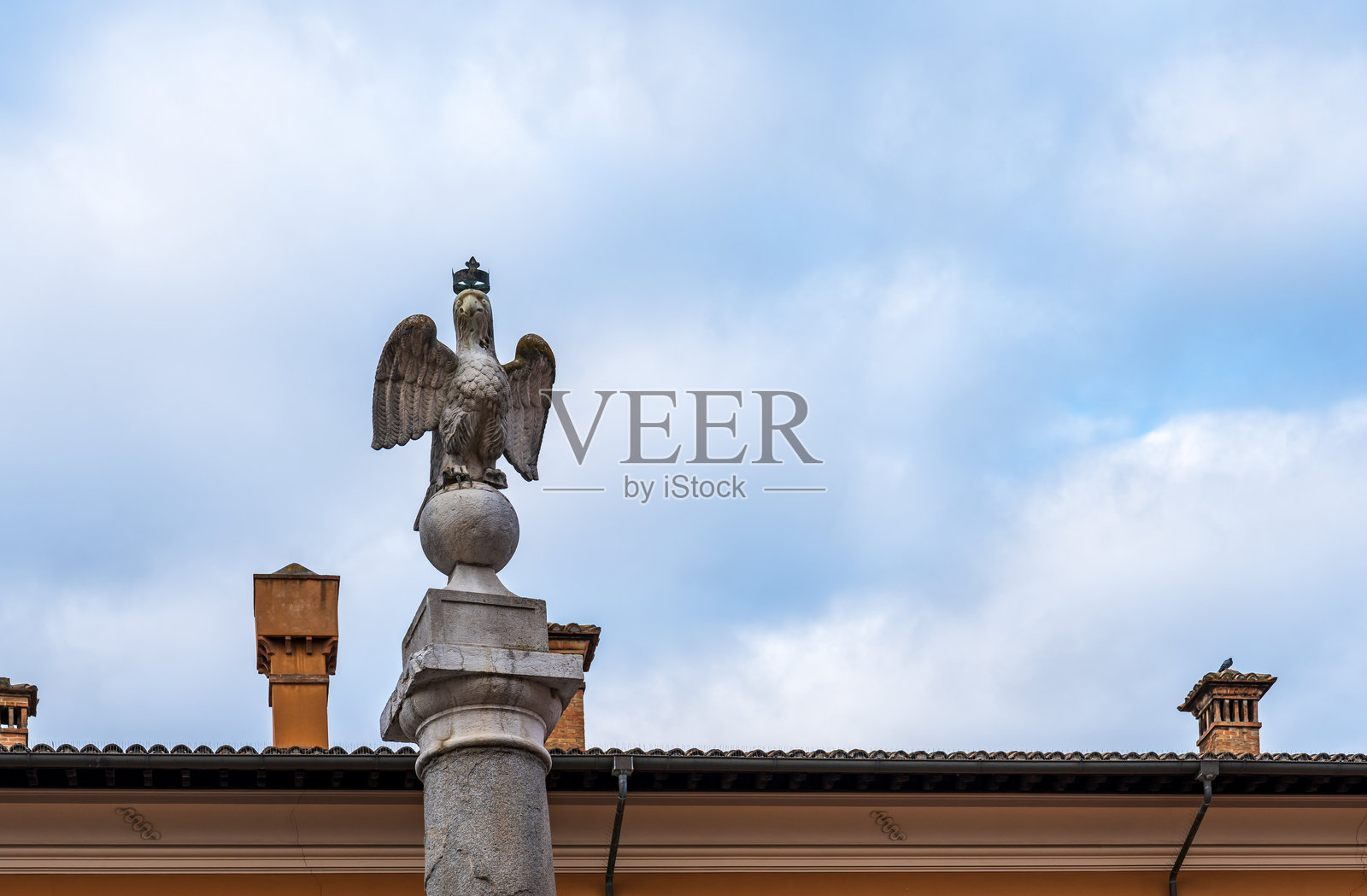 戴冠鹰栖息在大理石罗马柱顶上的地球上，背景是多云的天空和带烟囱的屋顶。照片摄影图片