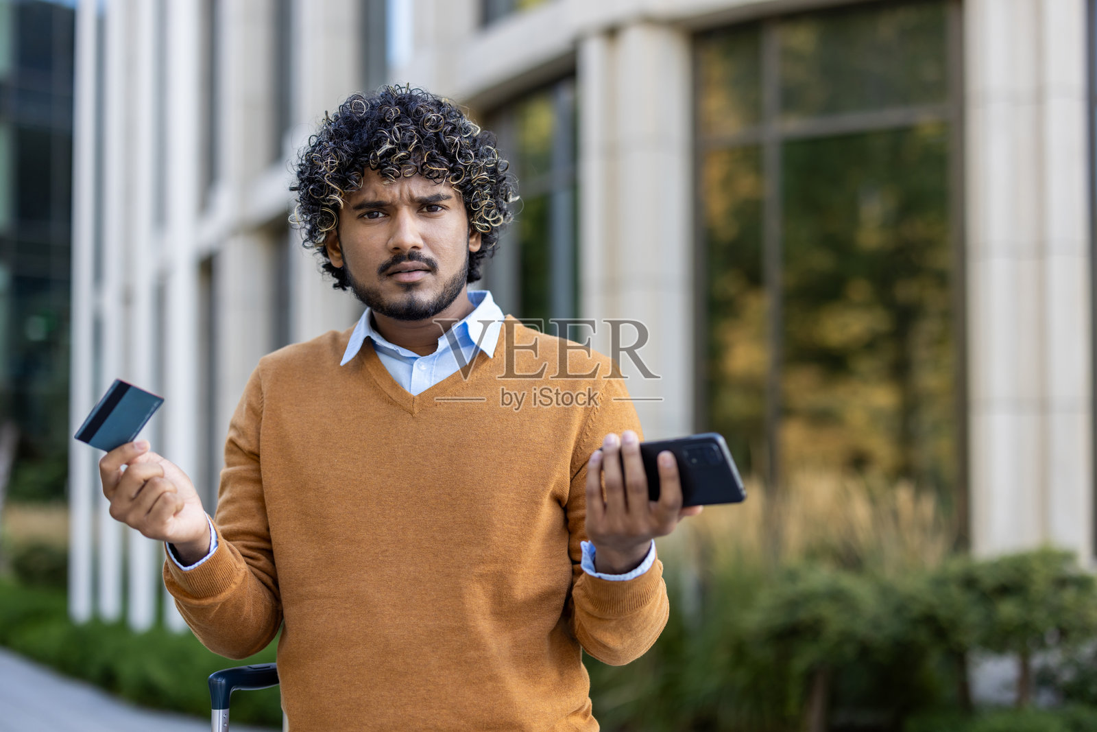 在现代办公楼外拿着信用卡和手机，看起来很担心的西班牙裔商人照片摄影图片