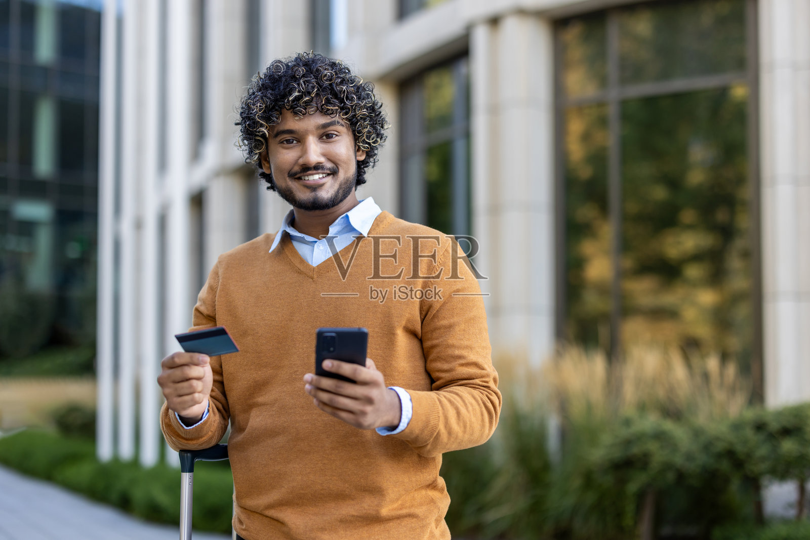 在现代建筑外，手持信用卡和手机的，穿着棕色毛衣的微笑男人，代表在线支付和商务旅行照片摄影图片