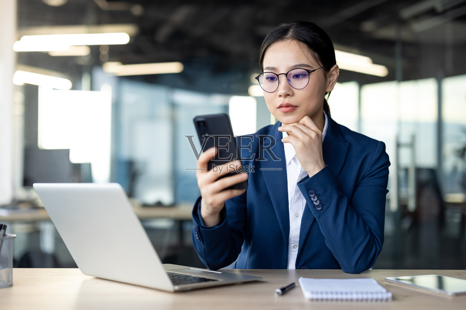 在办公室里使用智能手机，同时使用笔记本电脑工作的亚洲女商人，戴着眼镜和蓝色西装，专注于屏幕照片摄影图片