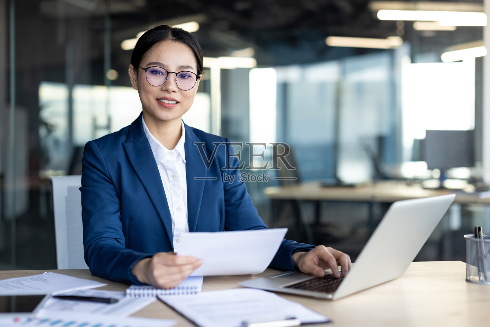 自信的亚洲女商人穿着蓝色西装，在现代办公室的办公桌前审查文件，配有笔记本电脑和文件照片摄影图片
