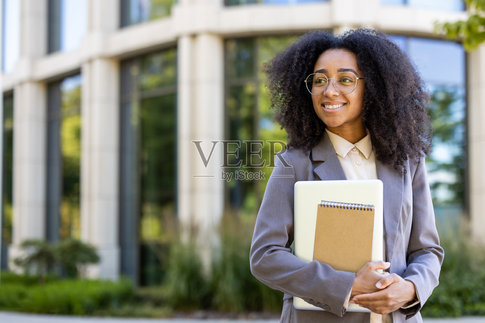 自信的年轻教师拿着笔记本和平板电脑在现代办公楼外，在自然光下微笑照片摄影图片