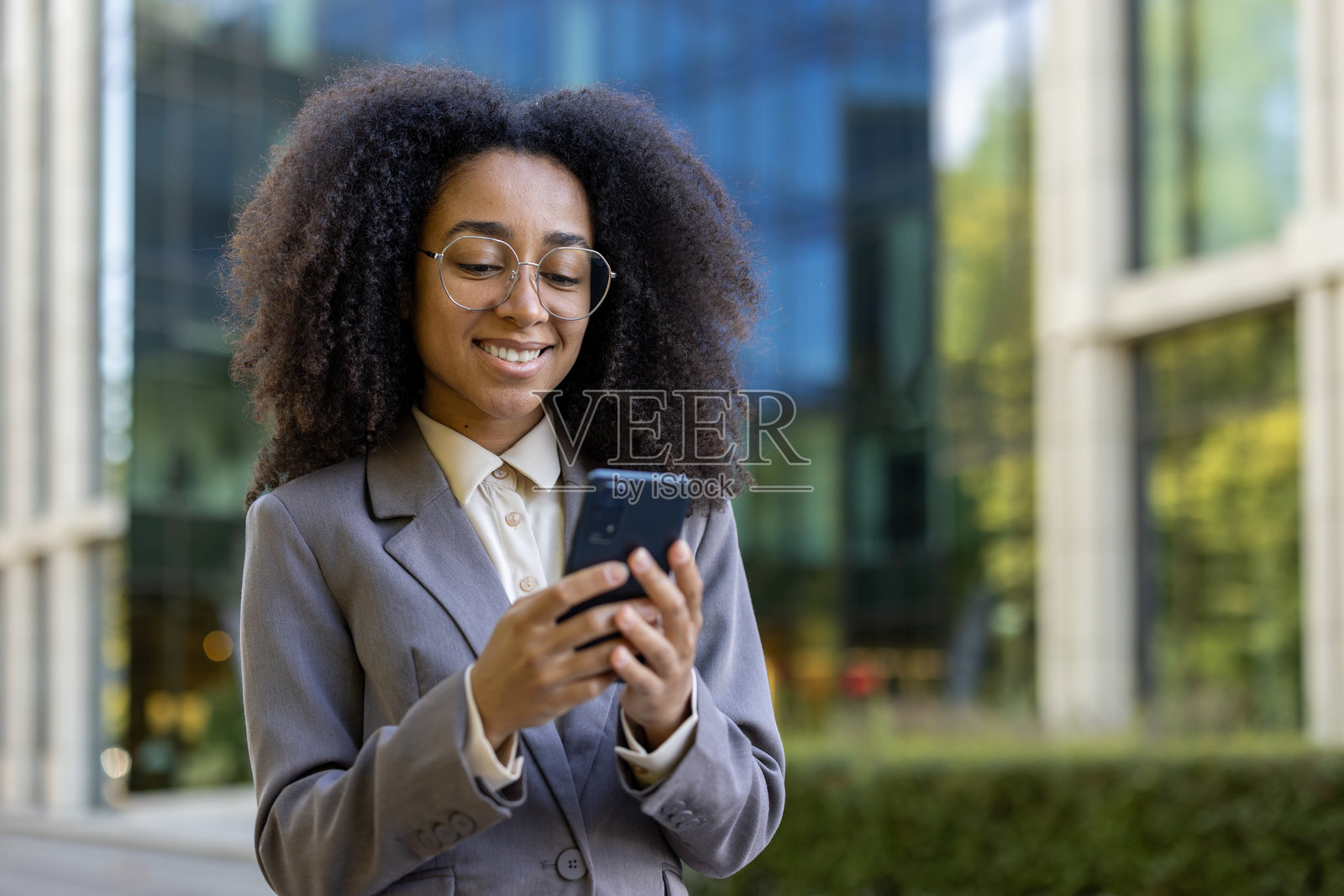 在现代办公楼外对着智能手机微笑的西班牙裔女商人，展现了专业环境中的自信和联系照片摄影图片