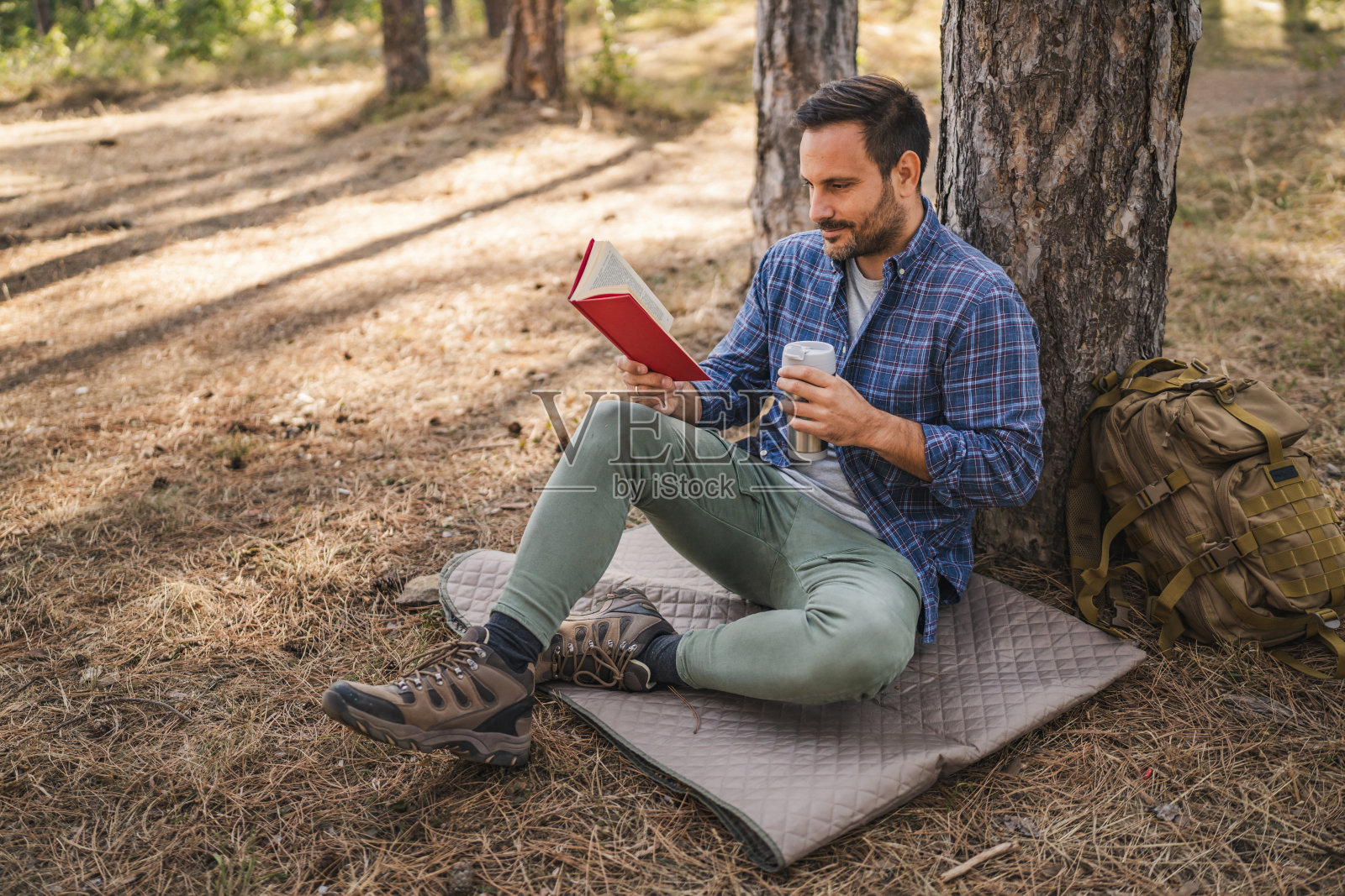 成年男人坐在树前的垫子上喝咖啡和看书照片摄影图片