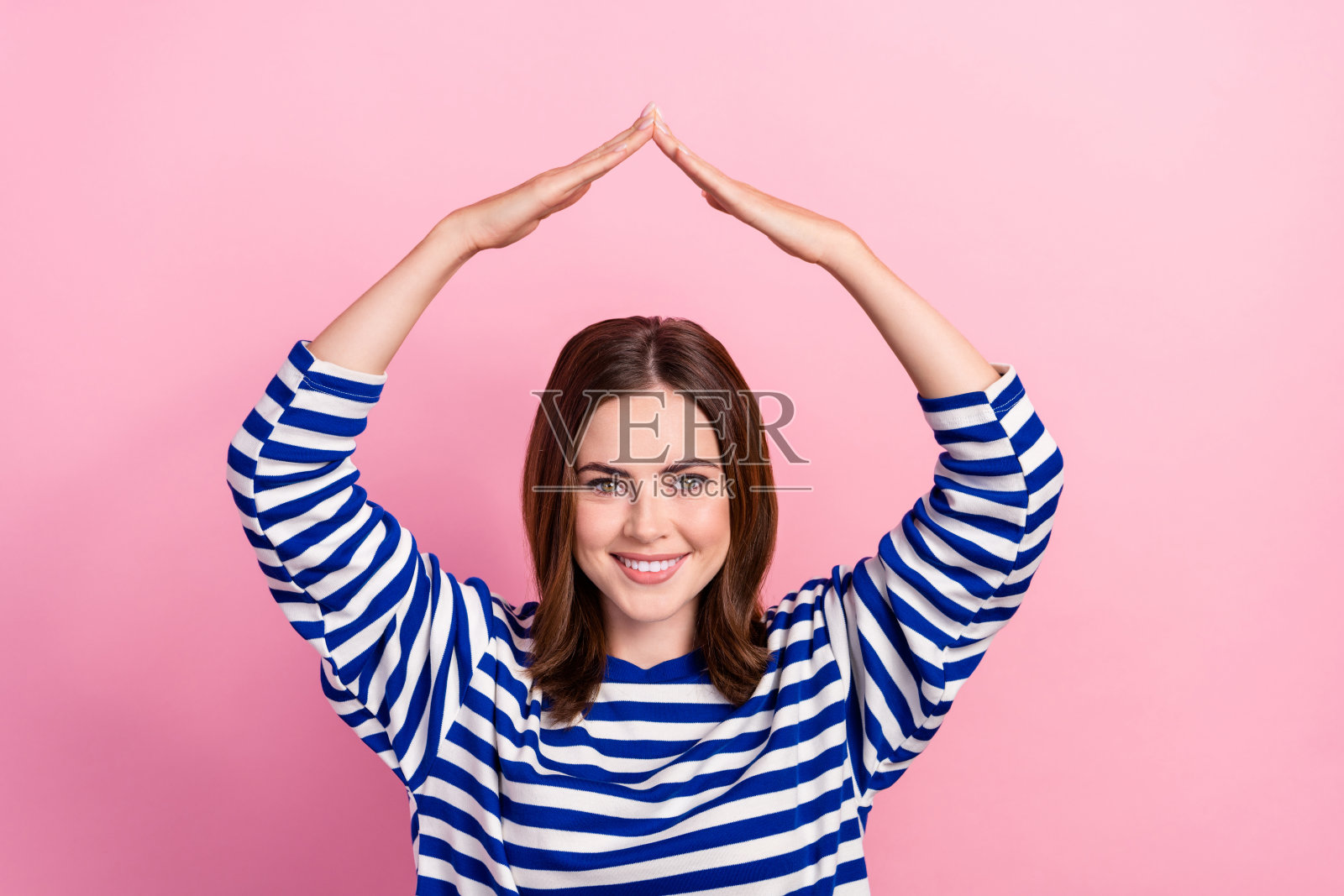 快乐漂亮女孩的肖像，露出牙齿的微笑，双臂在头顶做屋顶手势，背景为粉色。照片摄影图片