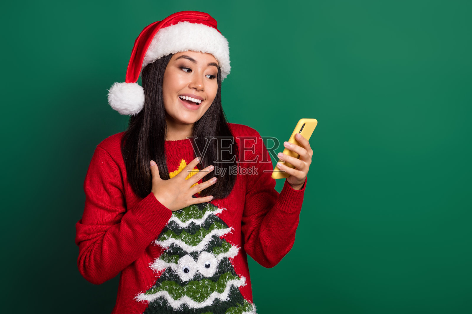 兴奋的搞笑女士穿着红色丑陋印花圣诞毛衣，戴着帽子，手持苹果和三星设备，背景为绿色，空白空间。照片摄影图片