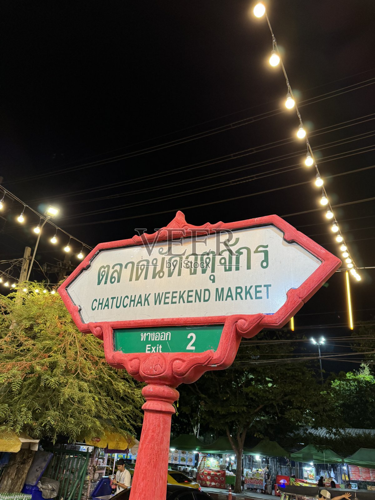 乍都乍周末市场红色柱子上的标志特写，夜间市场摊位灯火通明，人们在街头市场闲逛，前景聚焦照片摄影图片