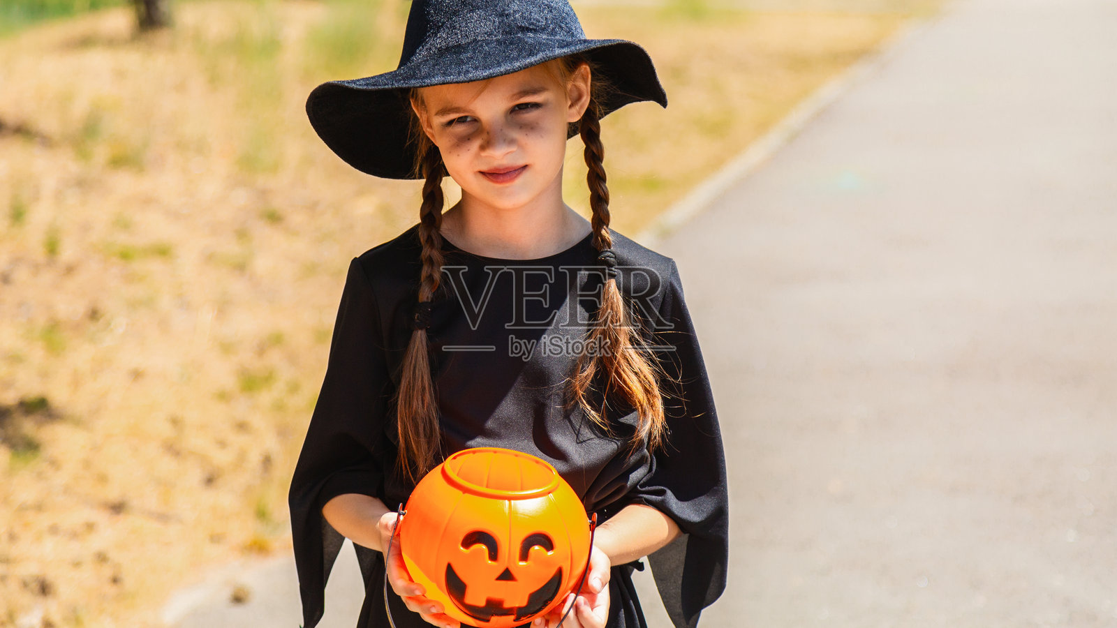 小女孩穿着女巫服装，手里拿着装满糖果和甜点的南瓜桶。孩子在万圣节期间进行“不给糖就捣蛋”。照片摄影图片