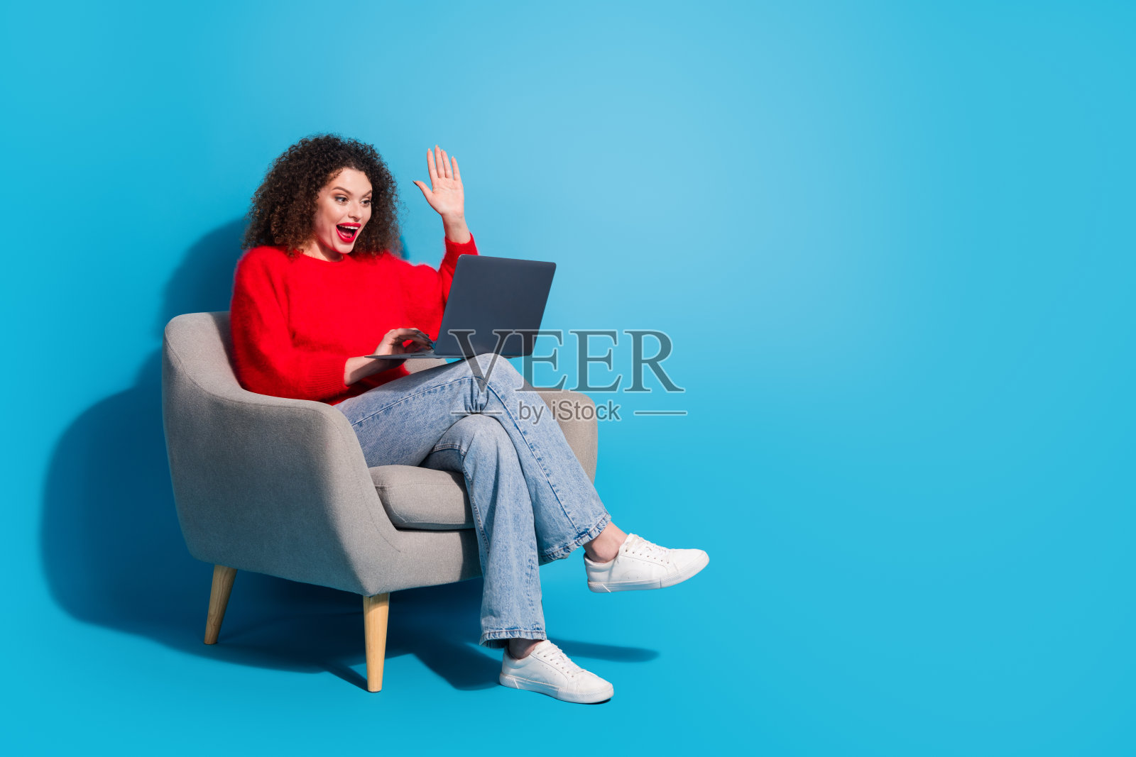 全身照，漂亮的年轻女孩坐在扶手椅上，手持上网本，挥手打招呼，穿着时尚的红色服装，背景为蓝色。照片摄影图片